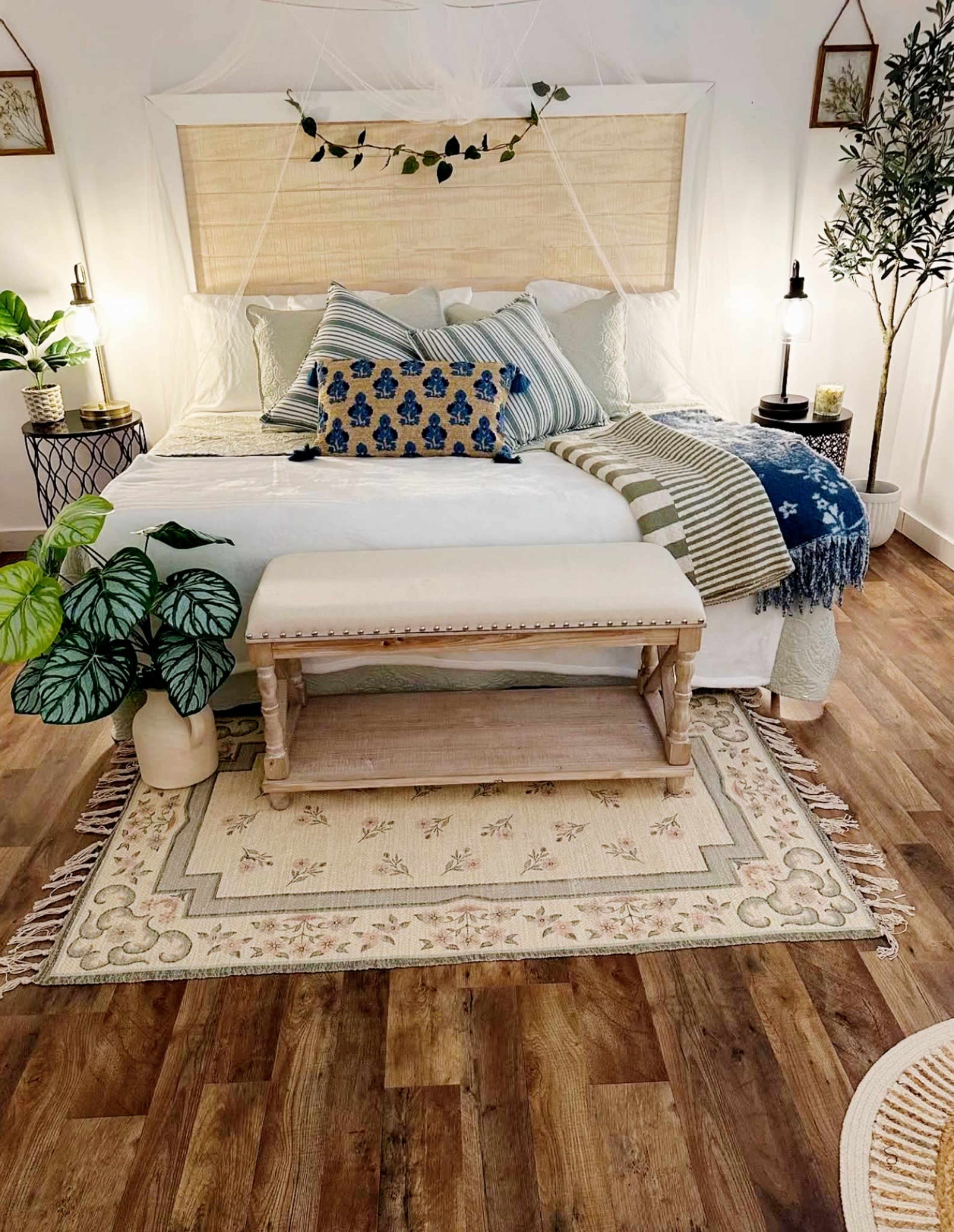 The image shows a neatly arranged bedroom featuring a large bed with decorative pillows, a bench at the foot, and plants surrounding a patterned rug on a wooden floor.