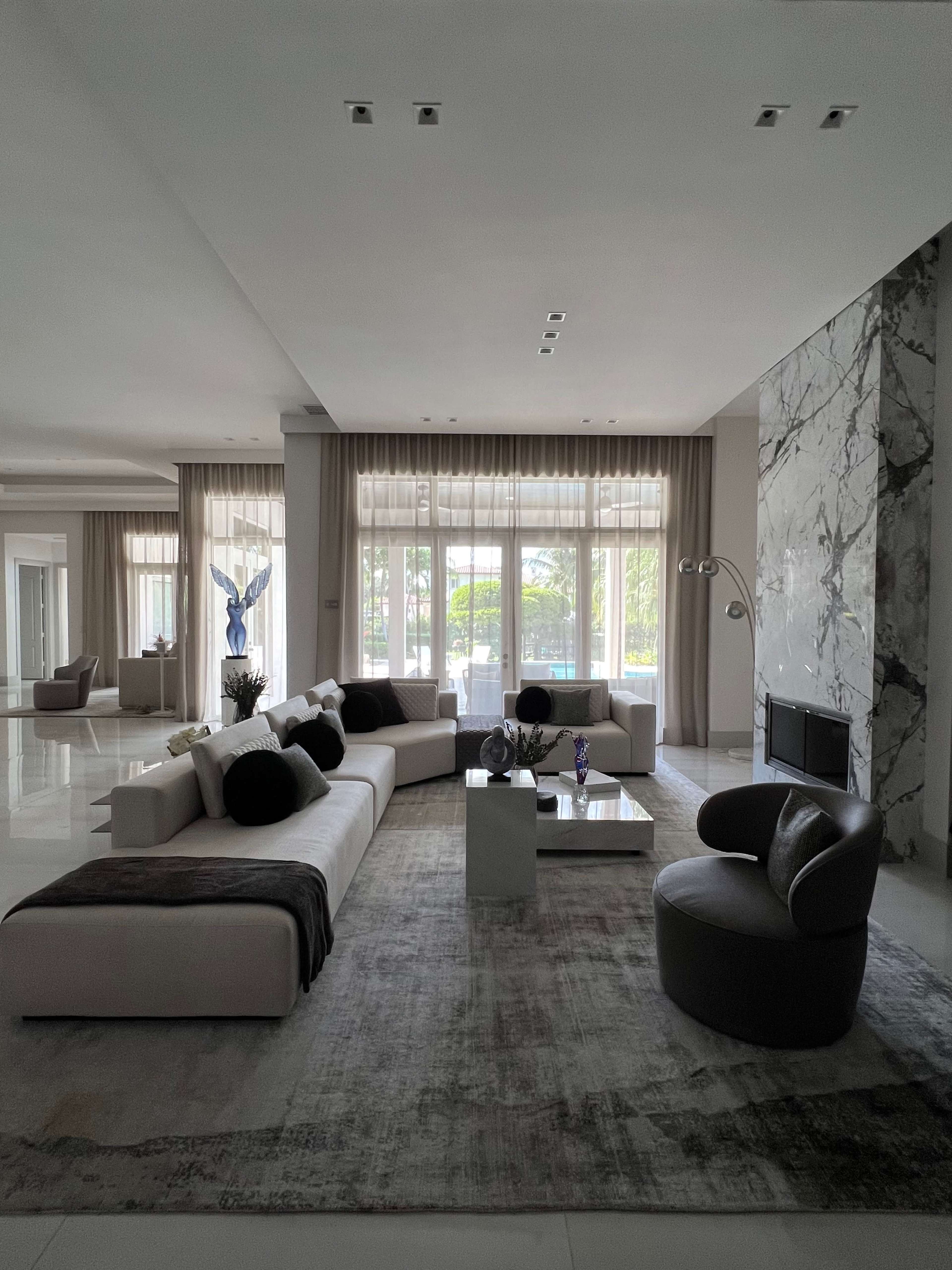 A spacious, modern living room features a beige sectional sofa, a round chair, and a coffee table on a light-colored rug, with large windows overlooking a patio.