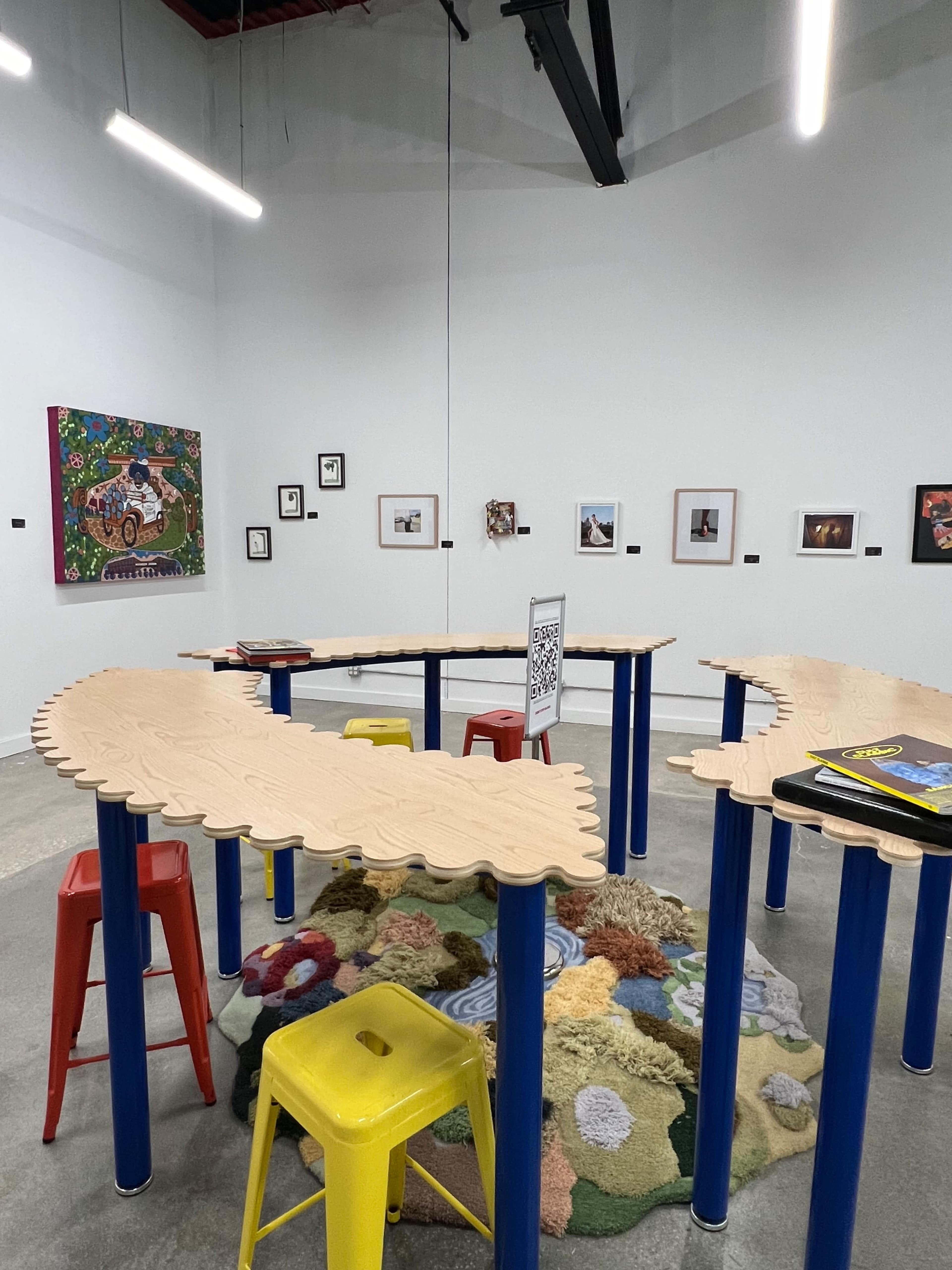 The image shows a modern art gallery featuring a curved wooden table surrounded by colorful stools, with various framed artworks displayed on the white walls.