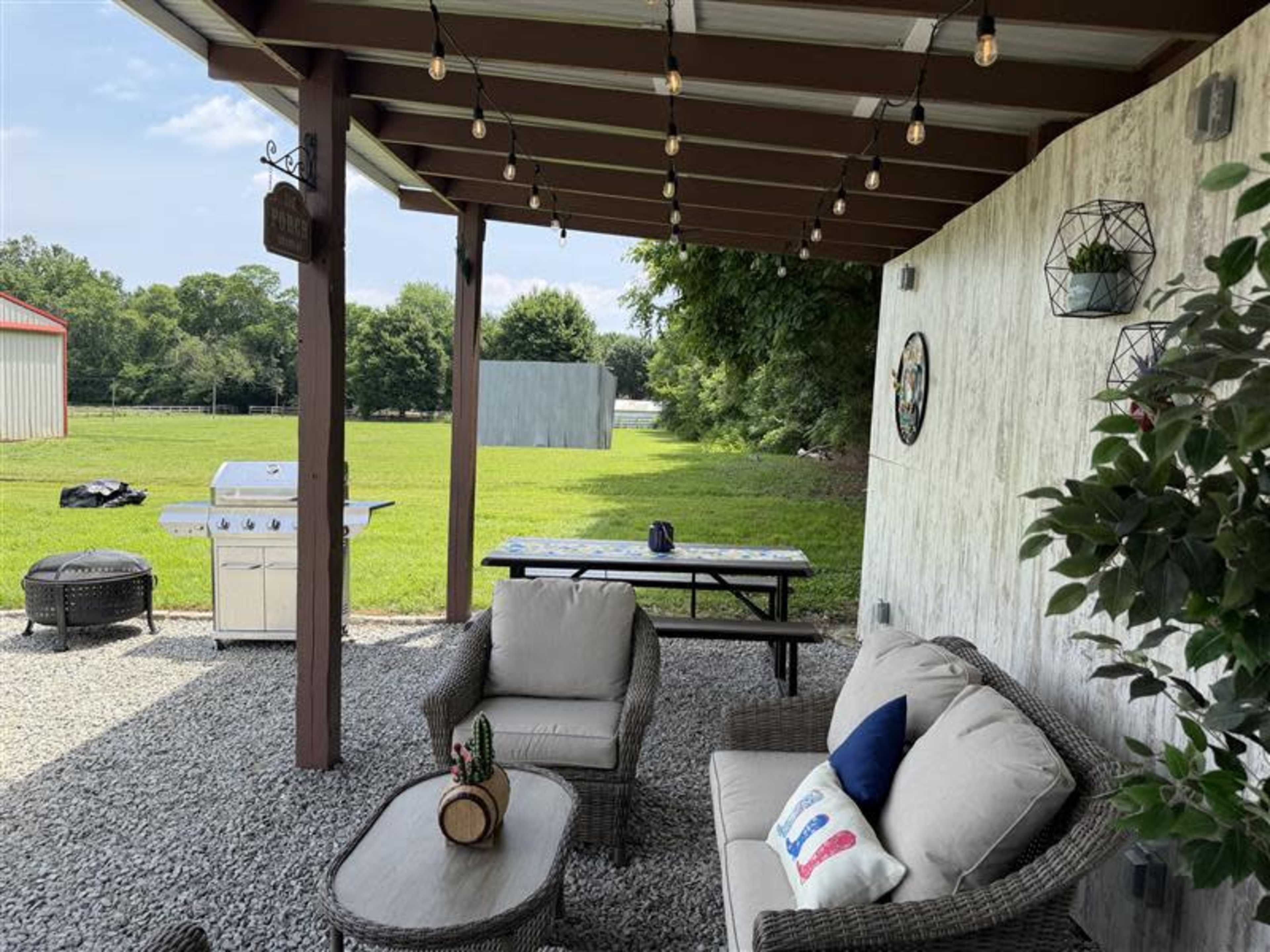 A covered patio area features comfortable seating, a grill, and a picnic table, surrounded by a grassy landscape.