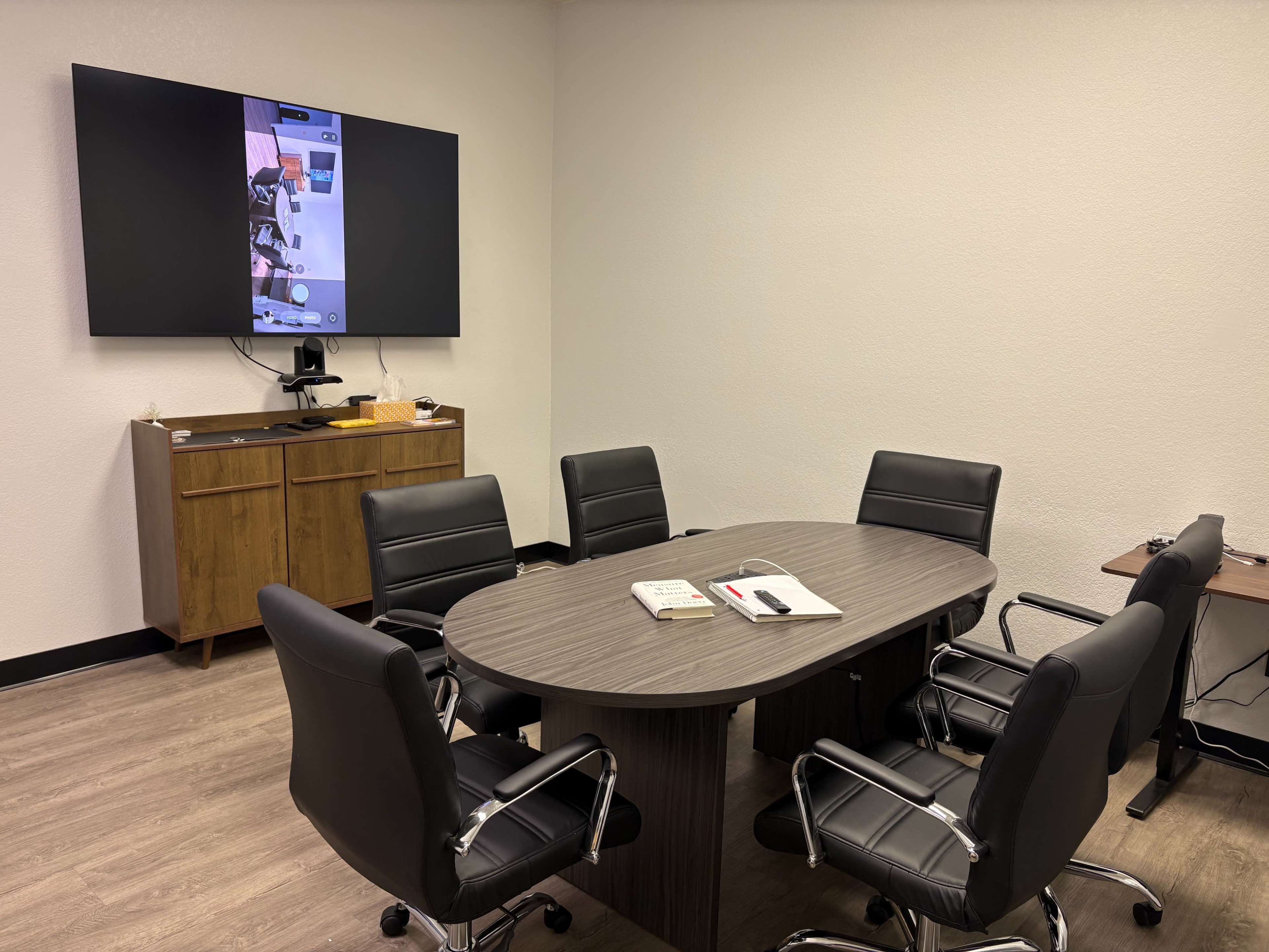 A conference room is equipped with a large table surrounded by six black chairs, and a screen displays content on the wall.