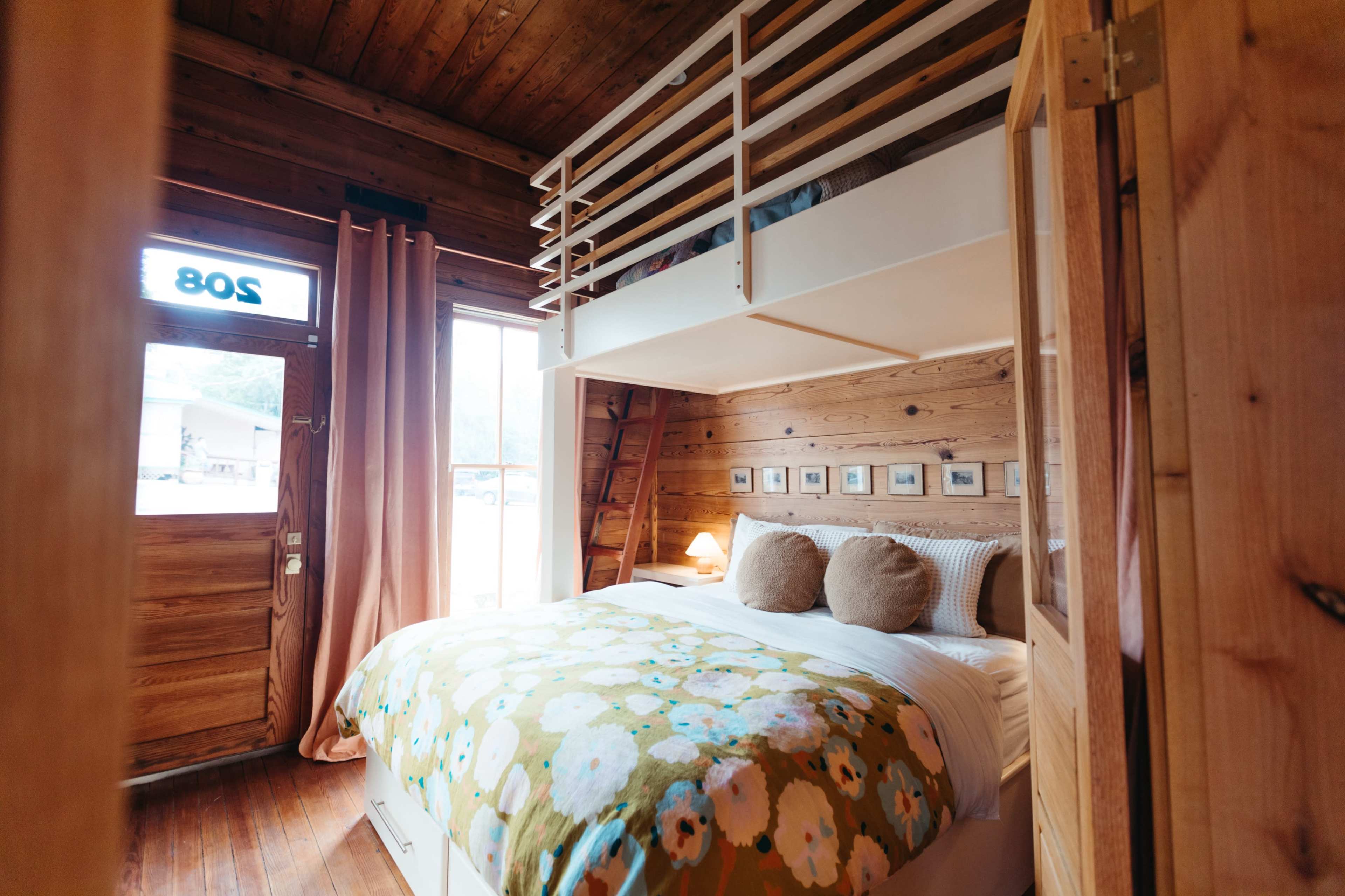 A cozy wooden bedroom featuring a double bed with a floral blanket, a loft area above, and a doorway with large windows.