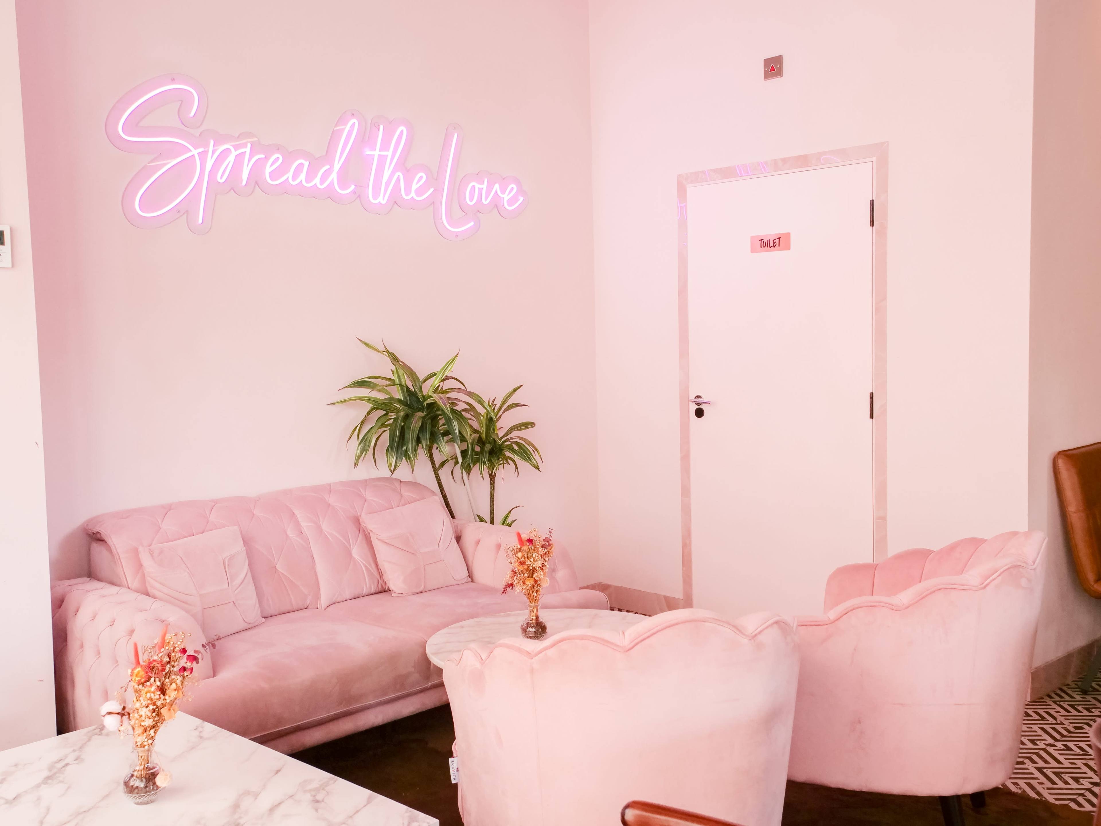 The image shows a pastel pink lounge area with plush seating, a plant, and a neon sign that reads "Spread the Love."