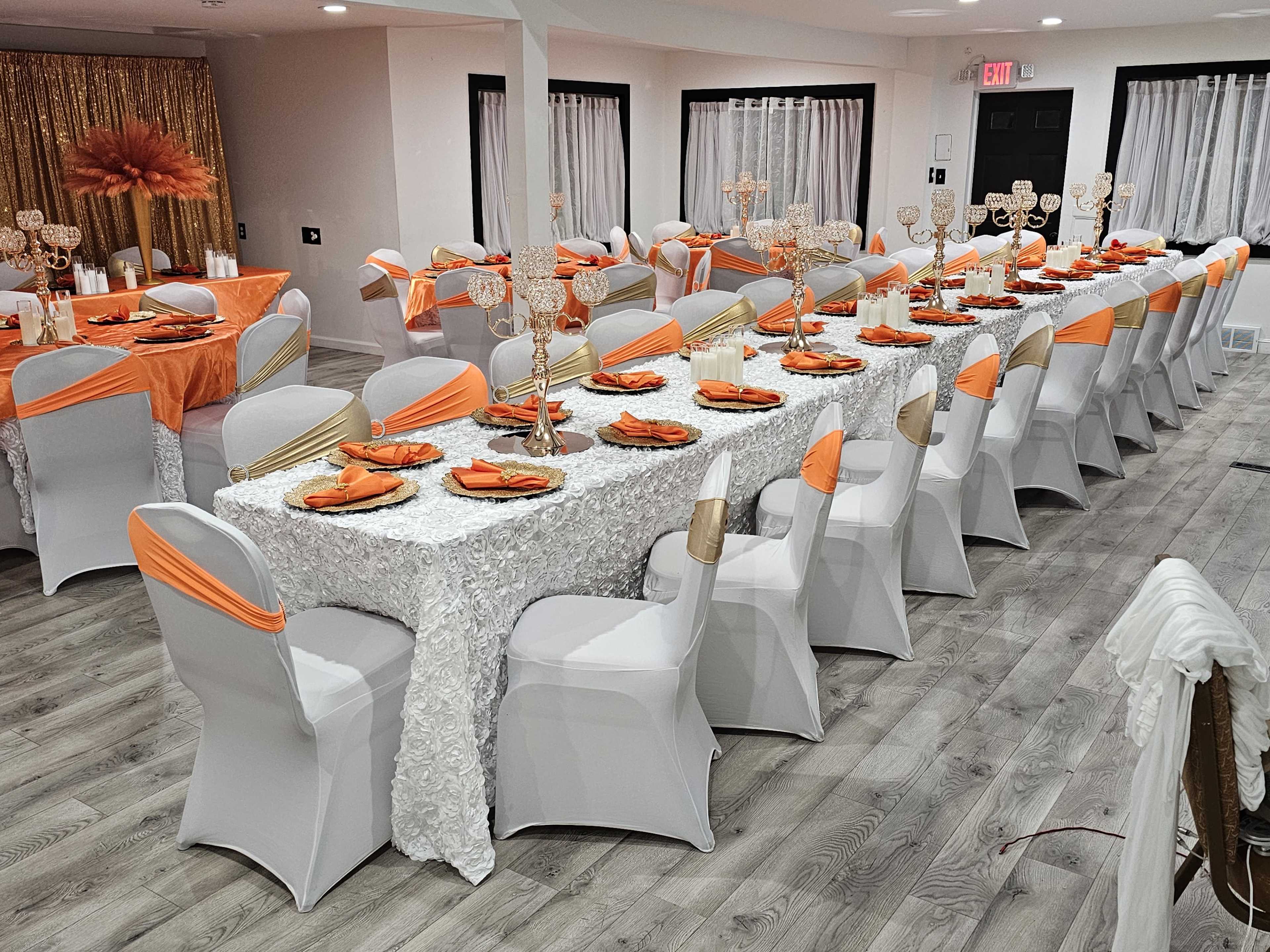 A long banquet table is set with white tablecloths and orange accents, surrounded by chairs draped in white with orange sashes, in a decorated dining area.