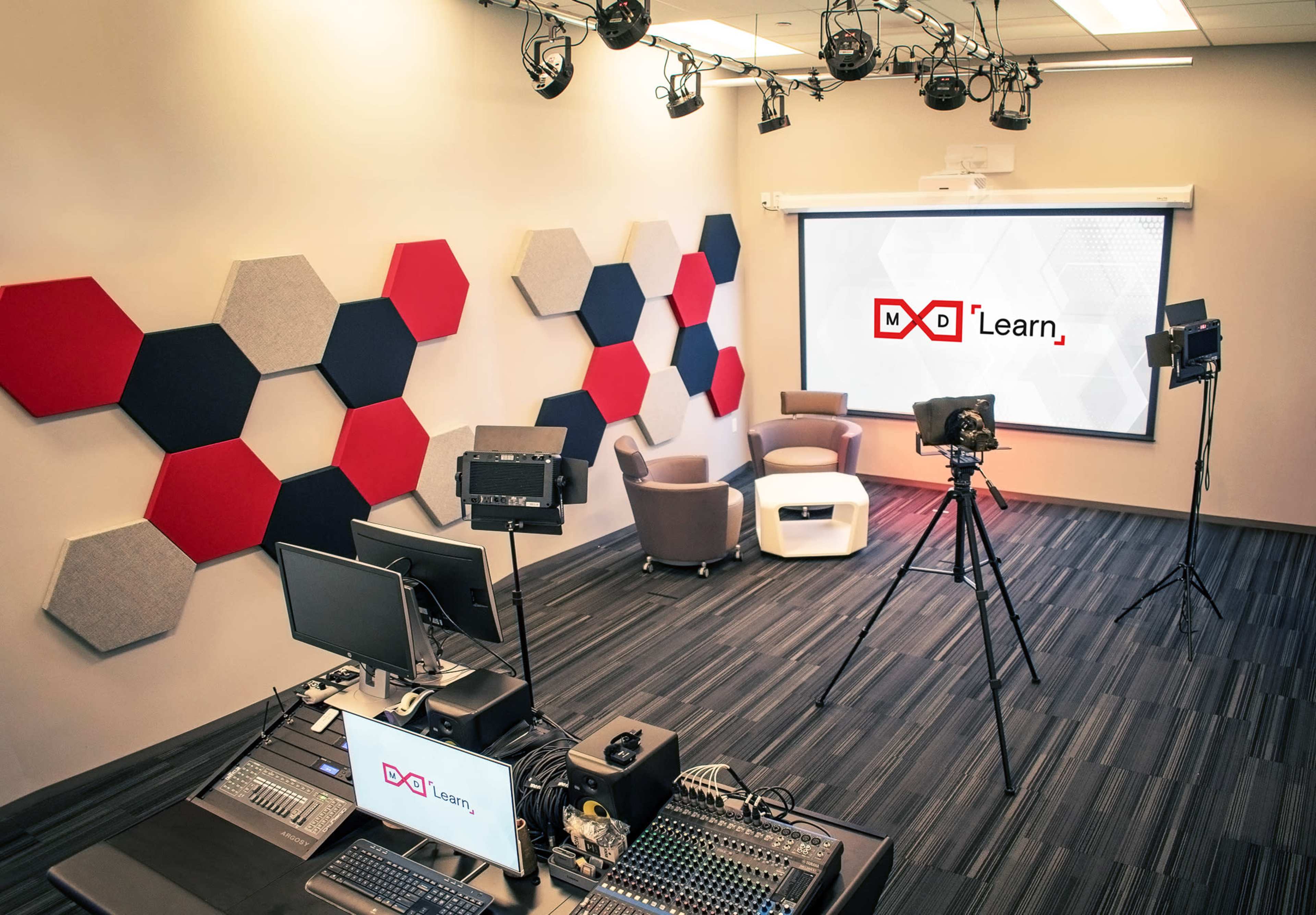 The image shows a modern studio setup featuring hexagonal wall decor, two chairs, and camera equipment ready for a video production.