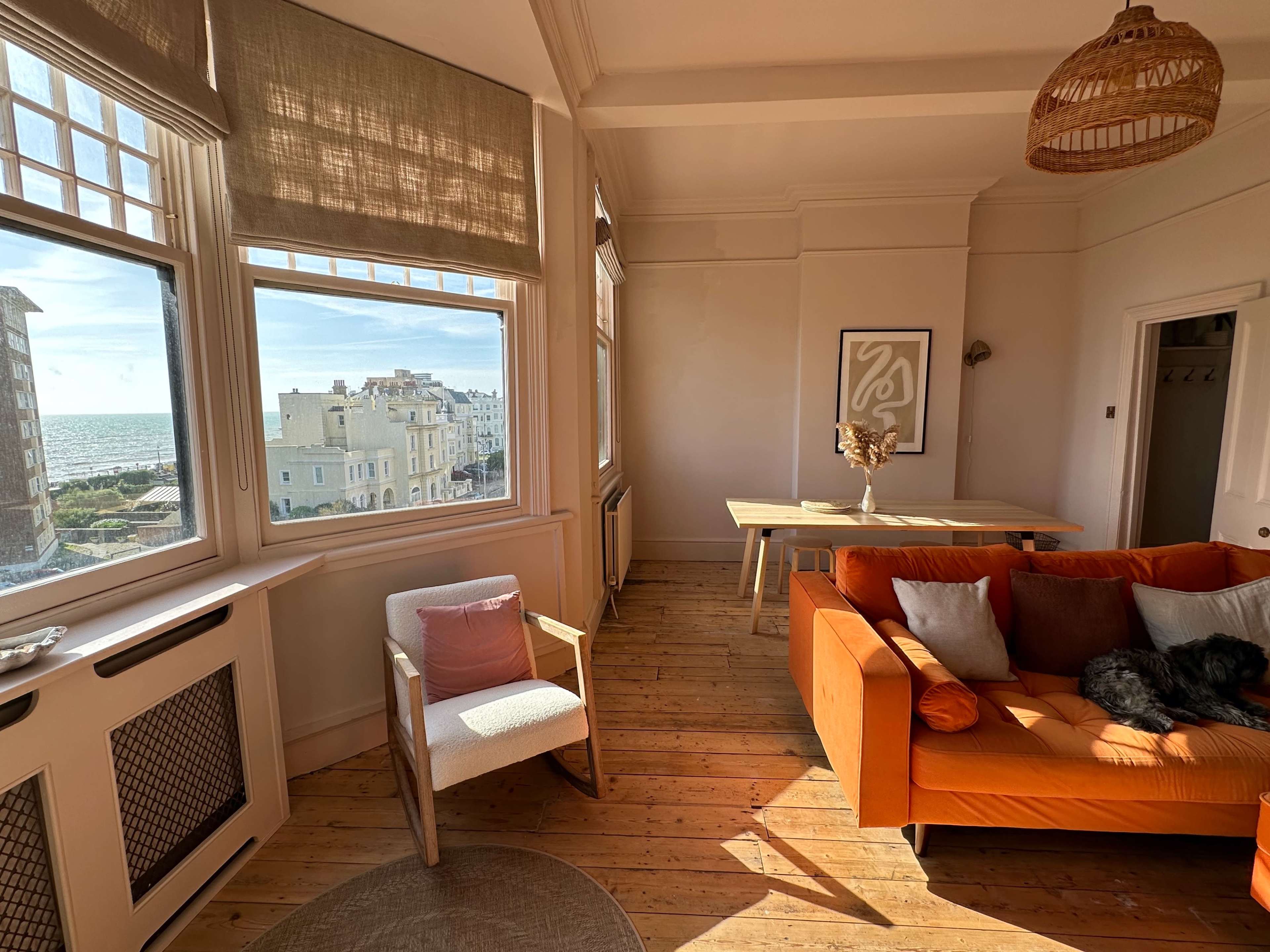 A bright interior with large windows overlooking a seaside view, featuring an orange couch, a wooden table, and a chair beside a circular rug on a wooden floor.
