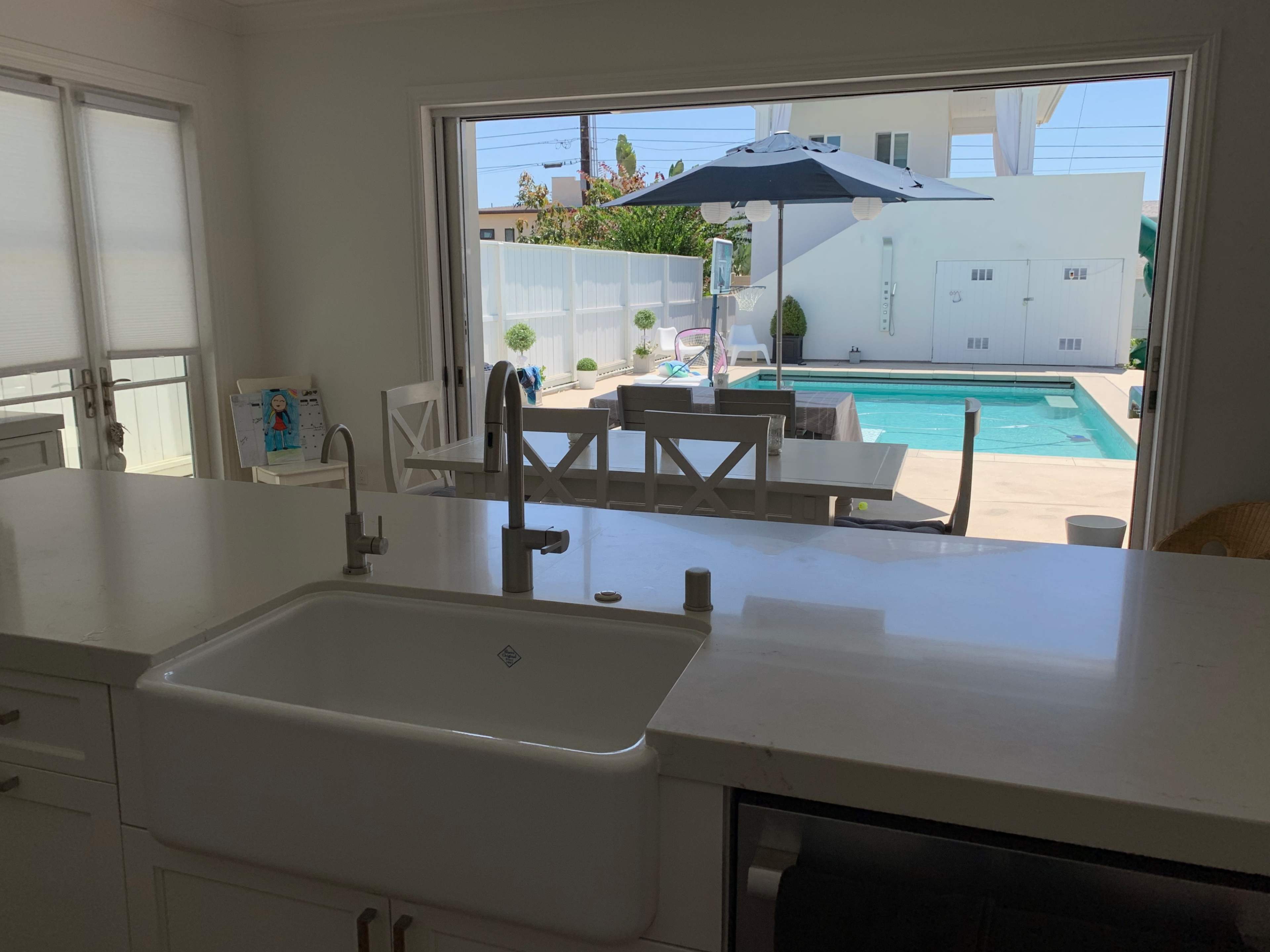 A kitchen with a farmhouse sink provides a view of a pool and outdoor seating area through a large open window.