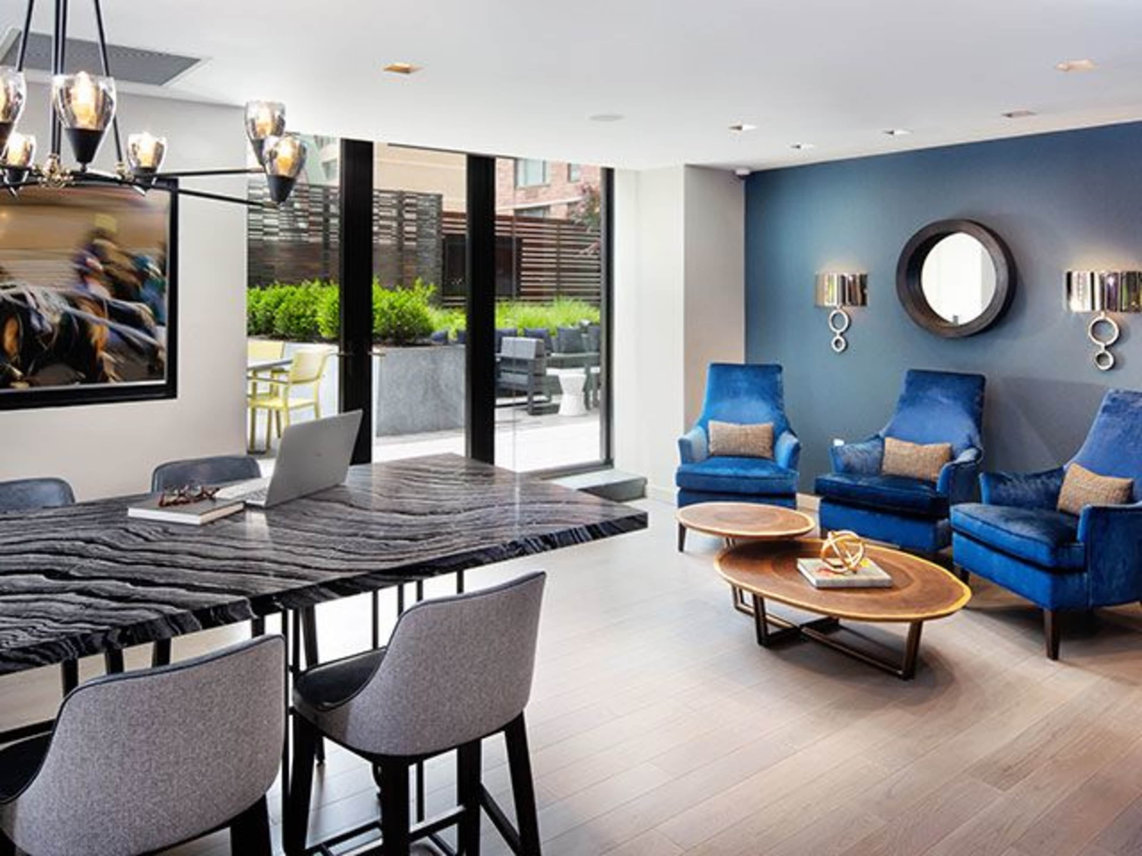 A modern living space features a large dining table, four chairs, and two blue armchairs arranged around a coffee table, with artwork and large windows overlooking a terrace.