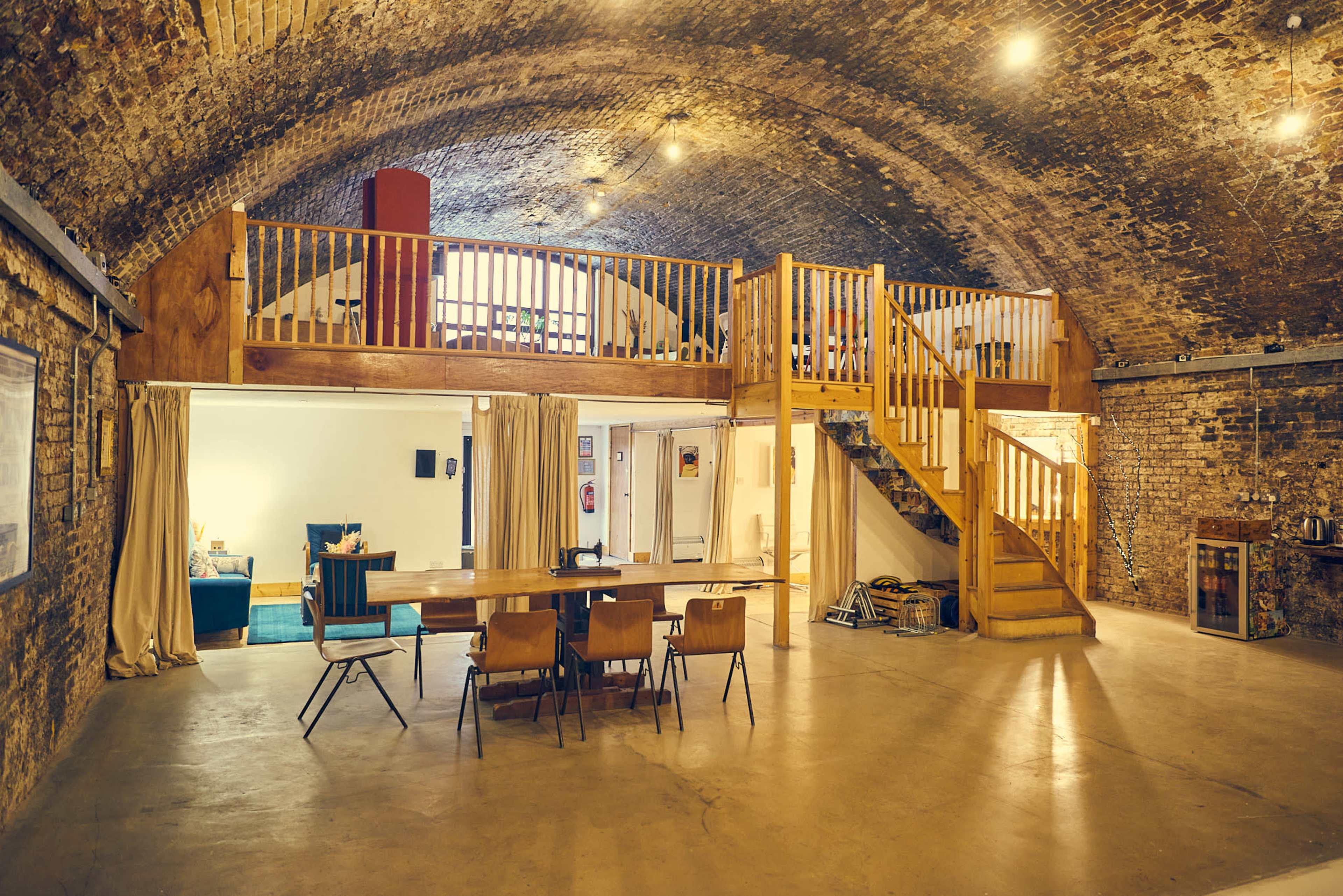 A spacious room with a wooden loft, a dining table with chairs, and a staircase leading to the upper level, all set within a brick archway structure.
