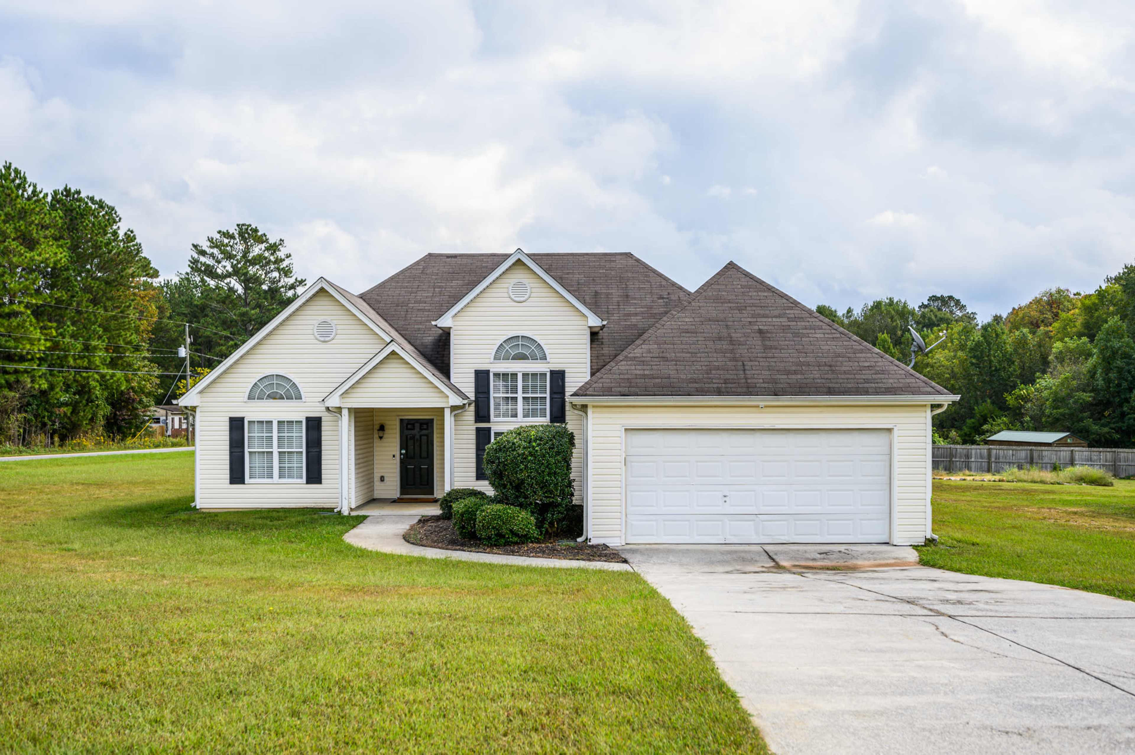 A single-story yellow house with a front porch and a two-car garage is situated on a grassy lot surrounded by trees.