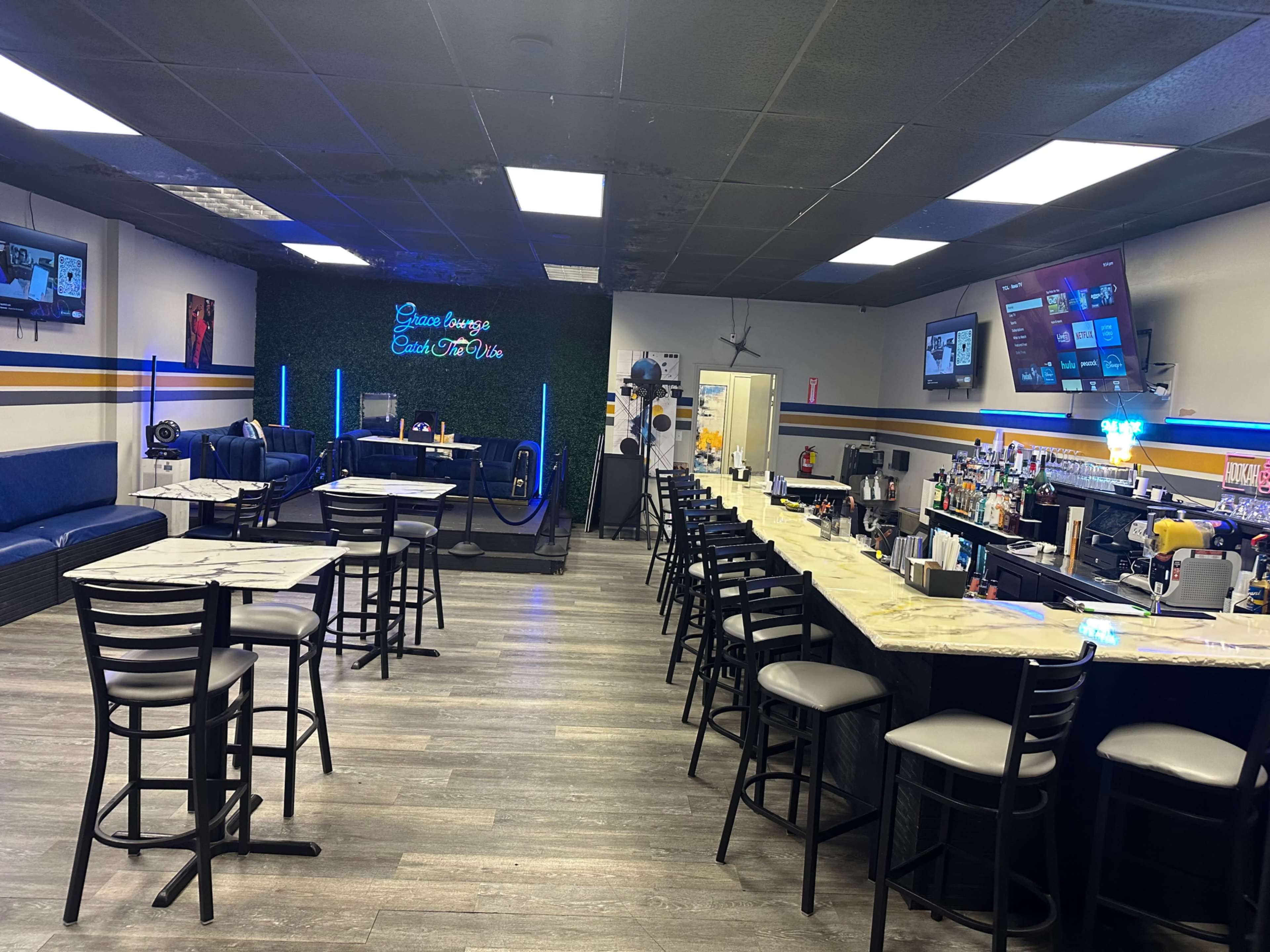 The image shows a modern bar and lounge area with a long counter, seating around tables, and two large screens on the walls.