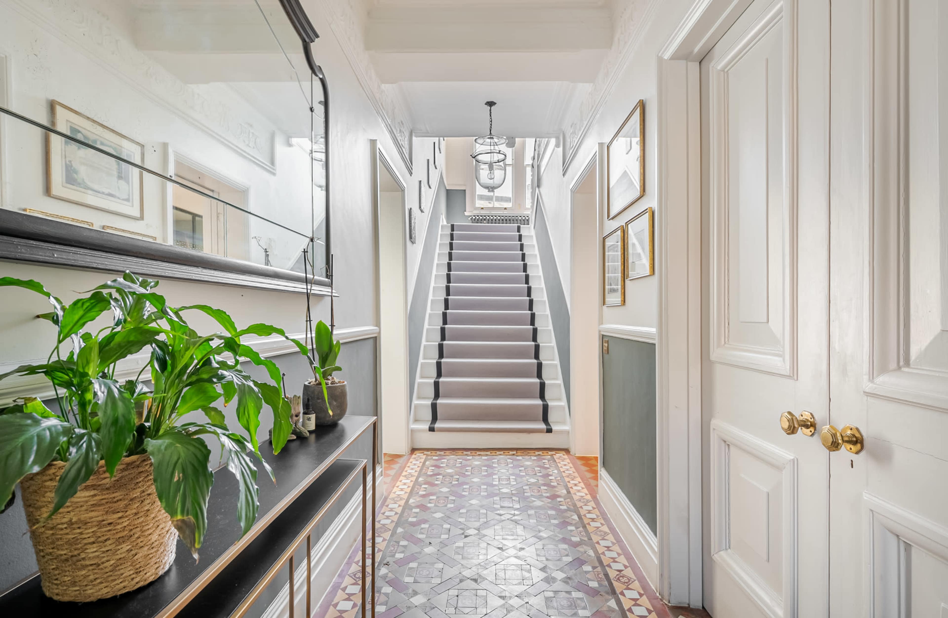 The image shows a hallway with a staircase at the end, featuring a plant on a side table and decorative items on the walls.