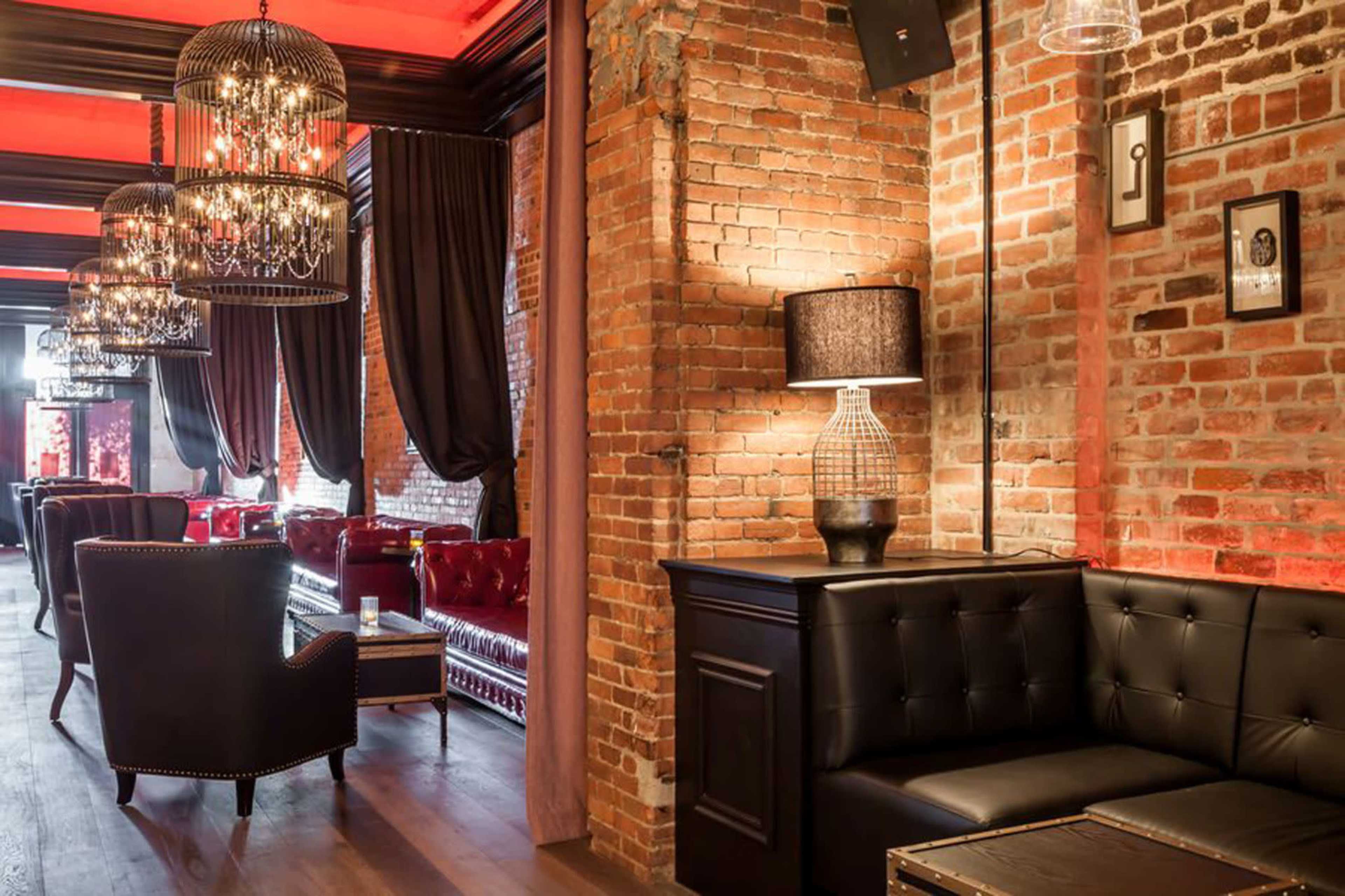 The image shows a stylish lounge with exposed brick walls, elegant chandeliers, plush red seating, and dark wood accents.