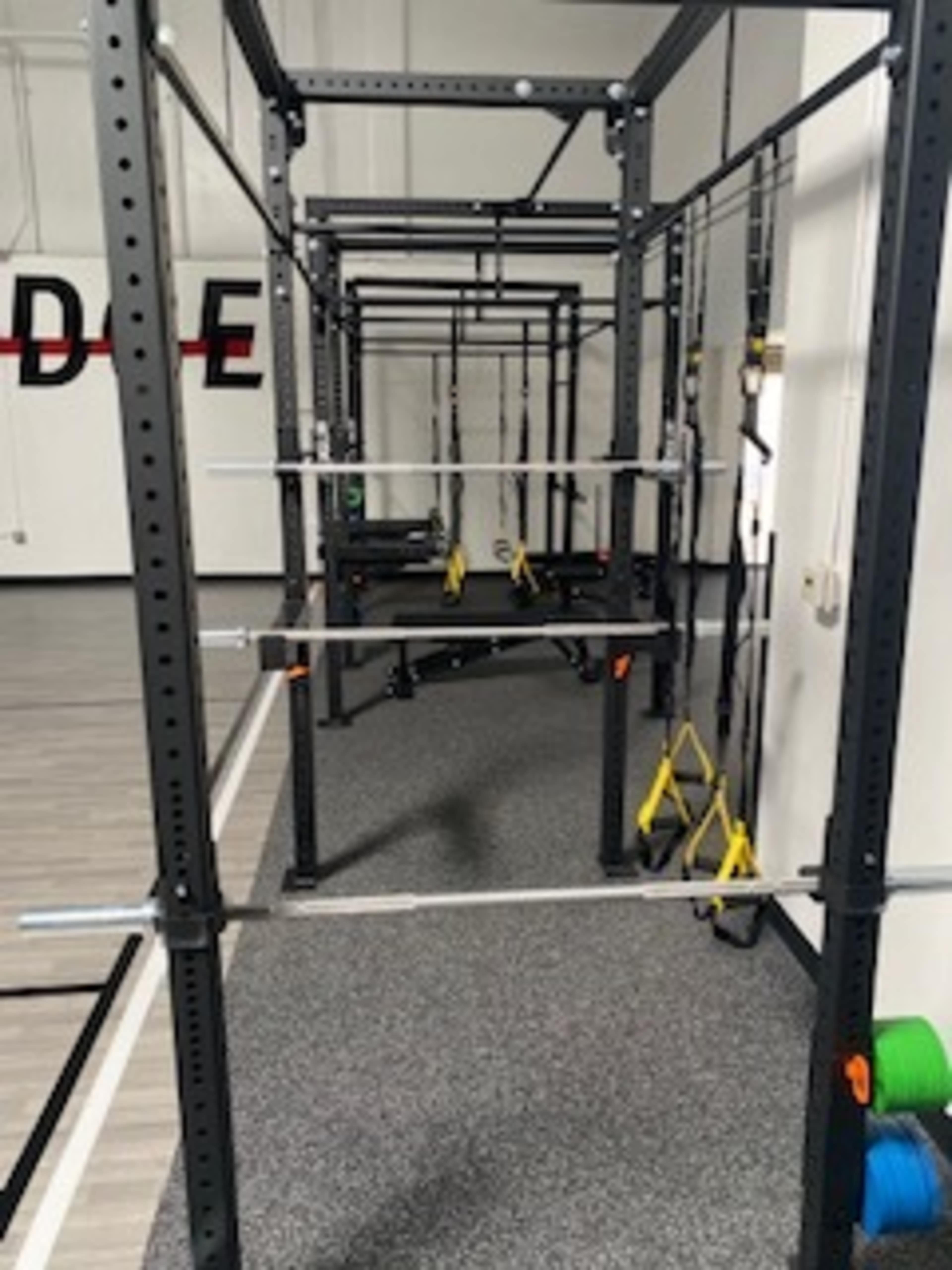 The image shows a gym area featuring a series of black squat racks and workout equipment set up on a gray flooring surface.