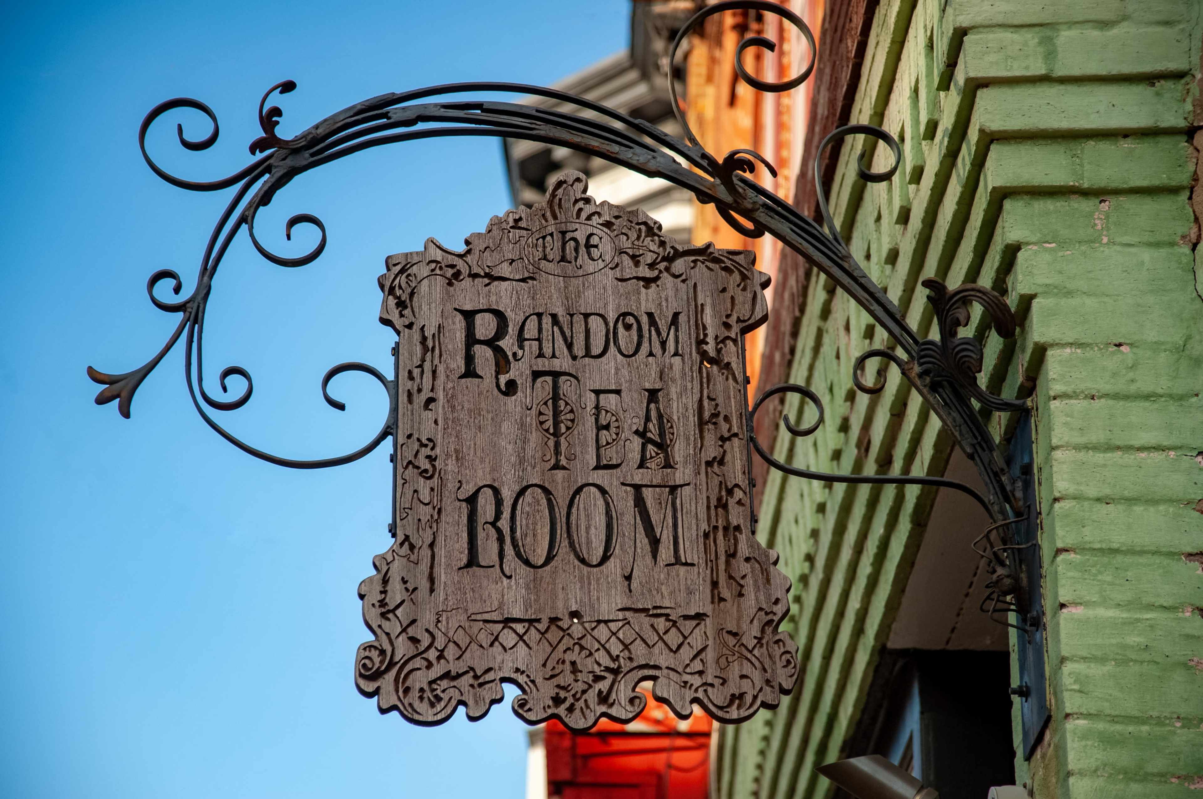 A wooden sign hanging from a wrought iron bracket reads "The Random Tea Room" against a clear blue sky.