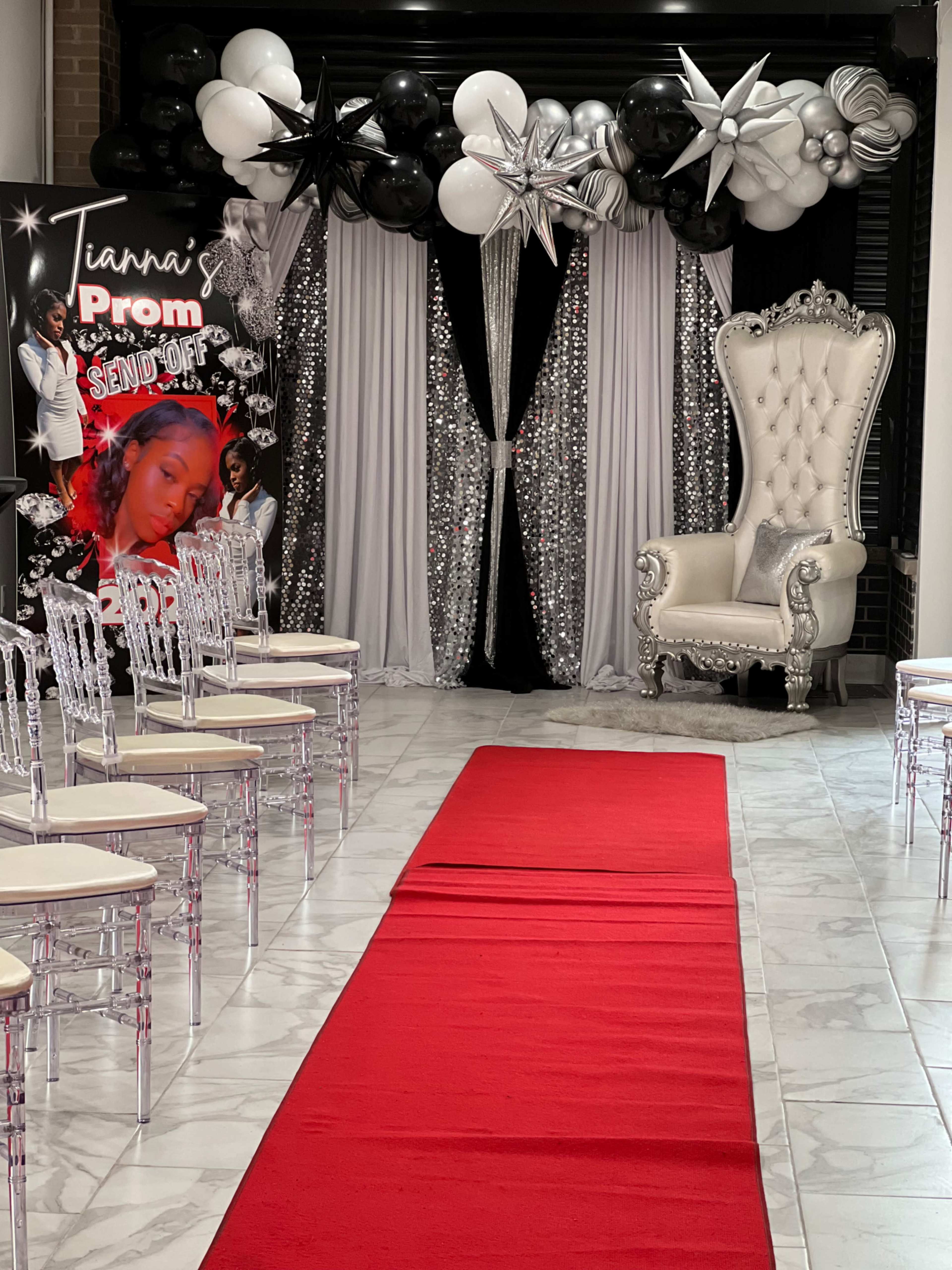 A prom setup featuring a red carpet leading to a decorative backdrop with balloons and stars, flanked by a luxurious throne and empty transparent chairs arranged along the sides.