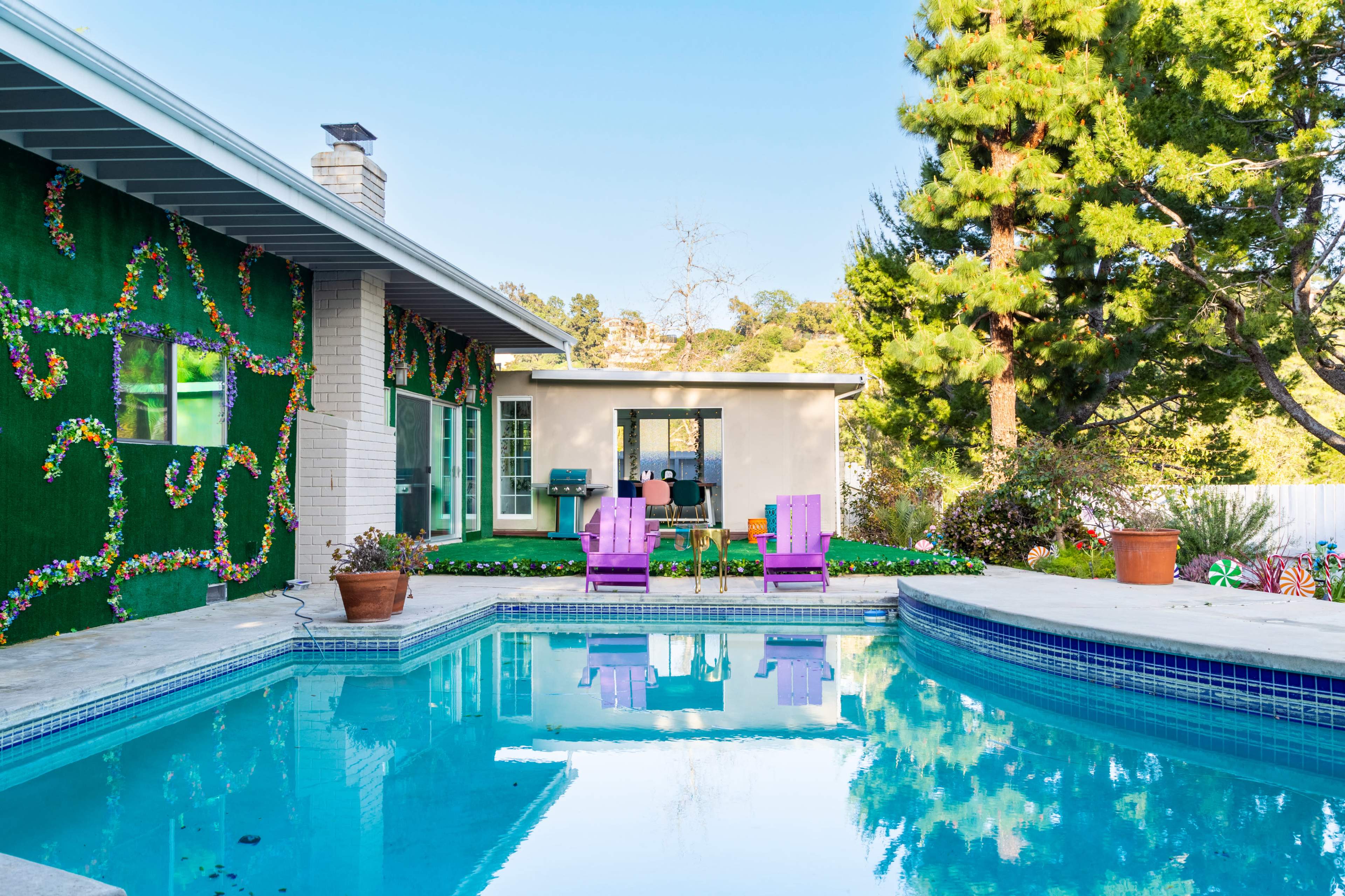 A clear pool reflects a colorful garden and a small outdoor seating area with purple chairs beside a modern house.