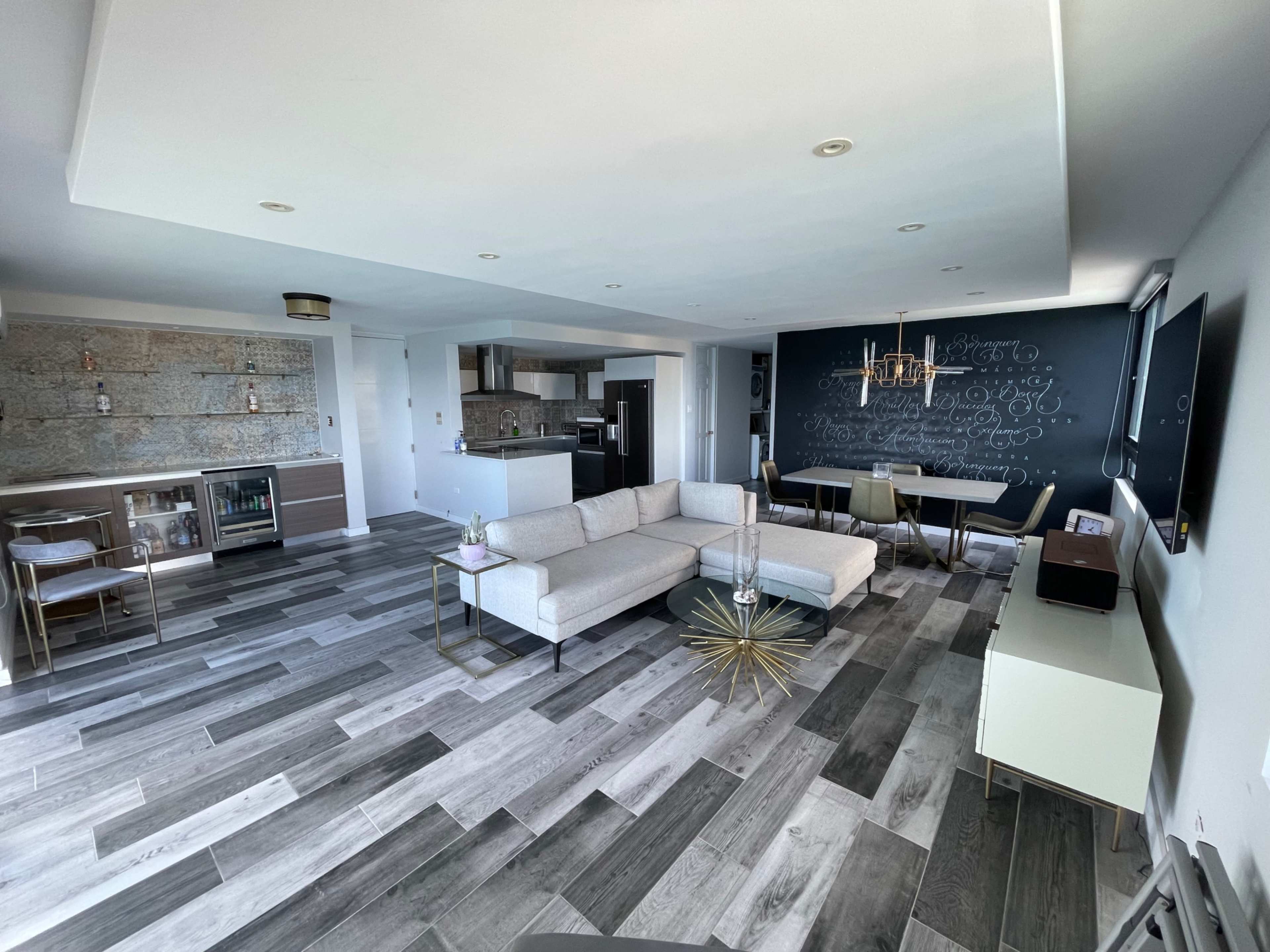 The image shows a contemporary living space featuring a seating area with a light-colored sectional sofa, a dining table, and a kitchen area with modern cabinetry and a bar setup.