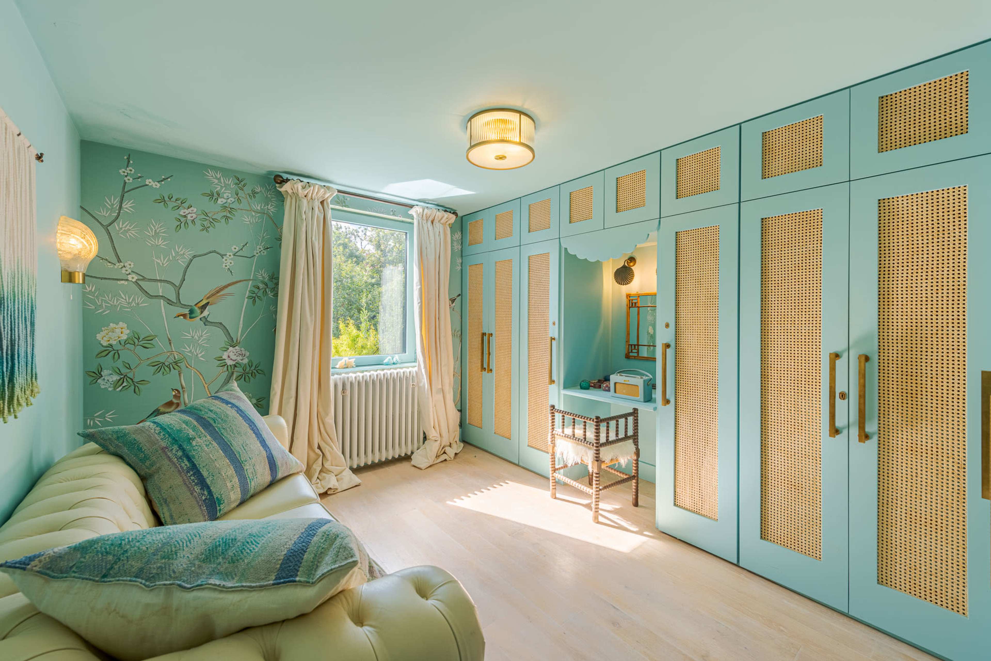 The image showcases a light blue room featuring a beige sofa, wicker-paneled wardrobes, and a small vanity table, all accented by floral wallpaper and large windows.