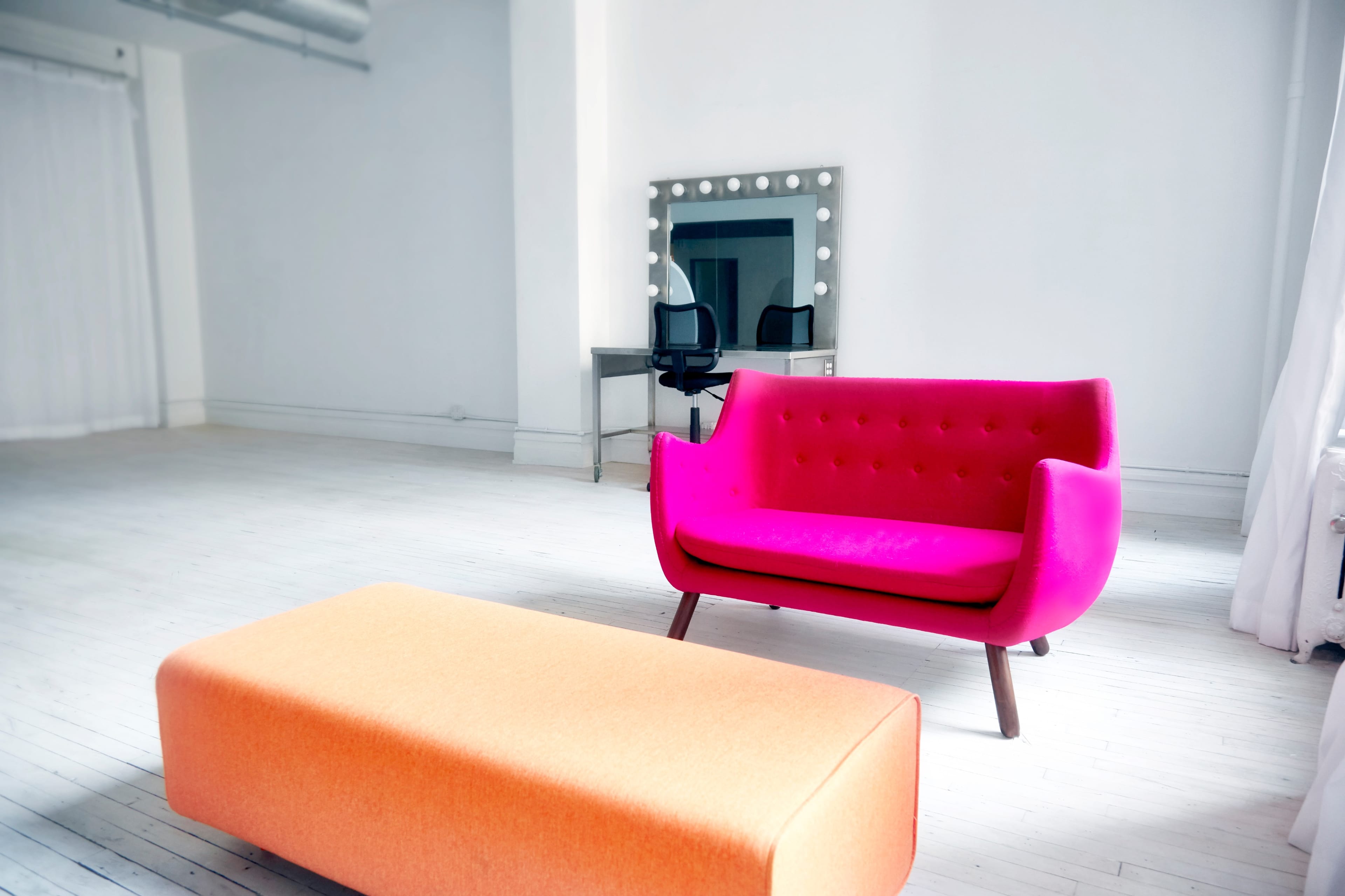 A bright pink sofa and an orange ottoman are positioned in a spacious, minimally decorated room with white walls and wooden floors.