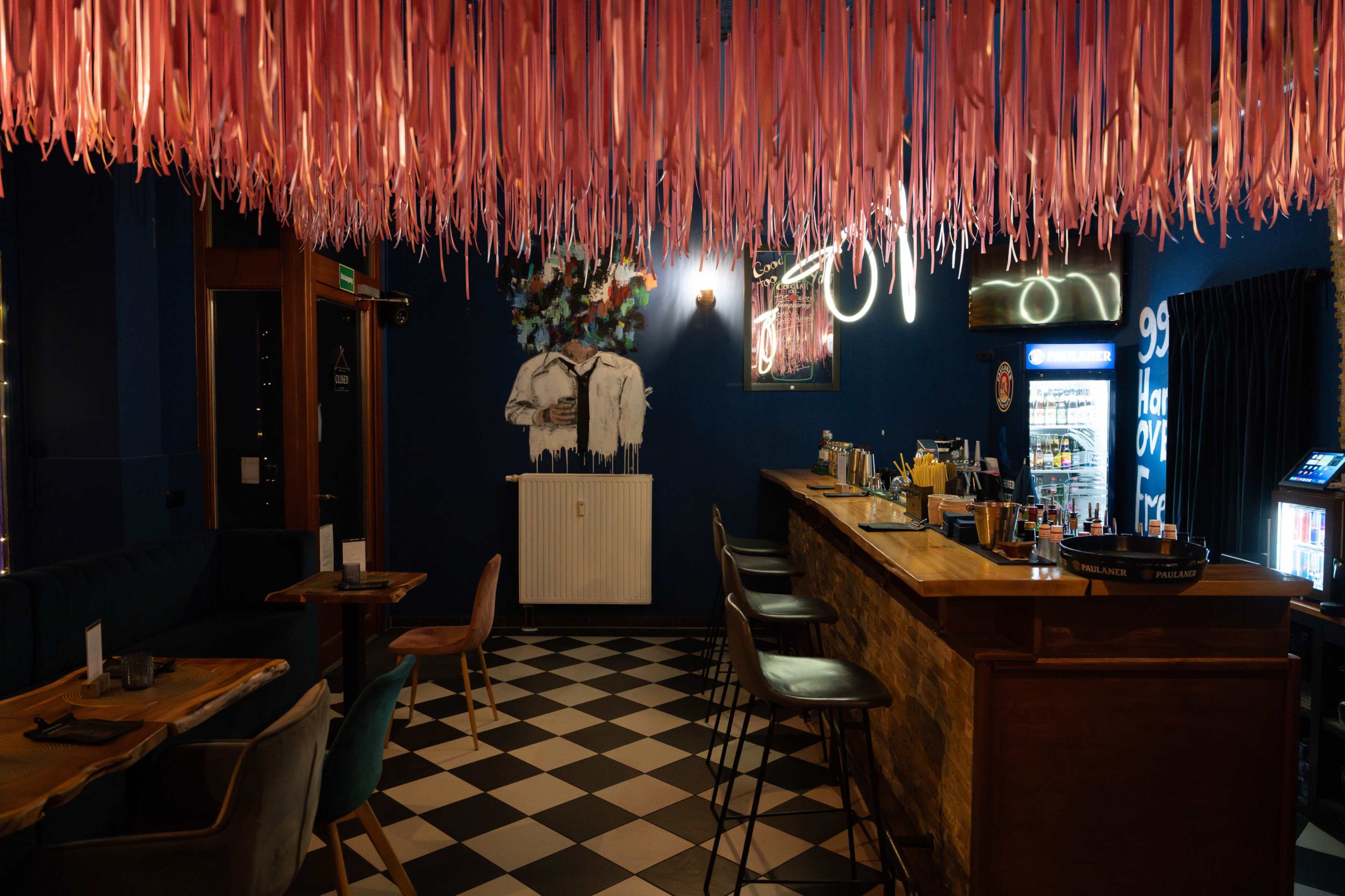 The image shows a stylish bar interior featuring pink streamers hanging from the ceiling, a wooden bar with various drinks, and checkered flooring.