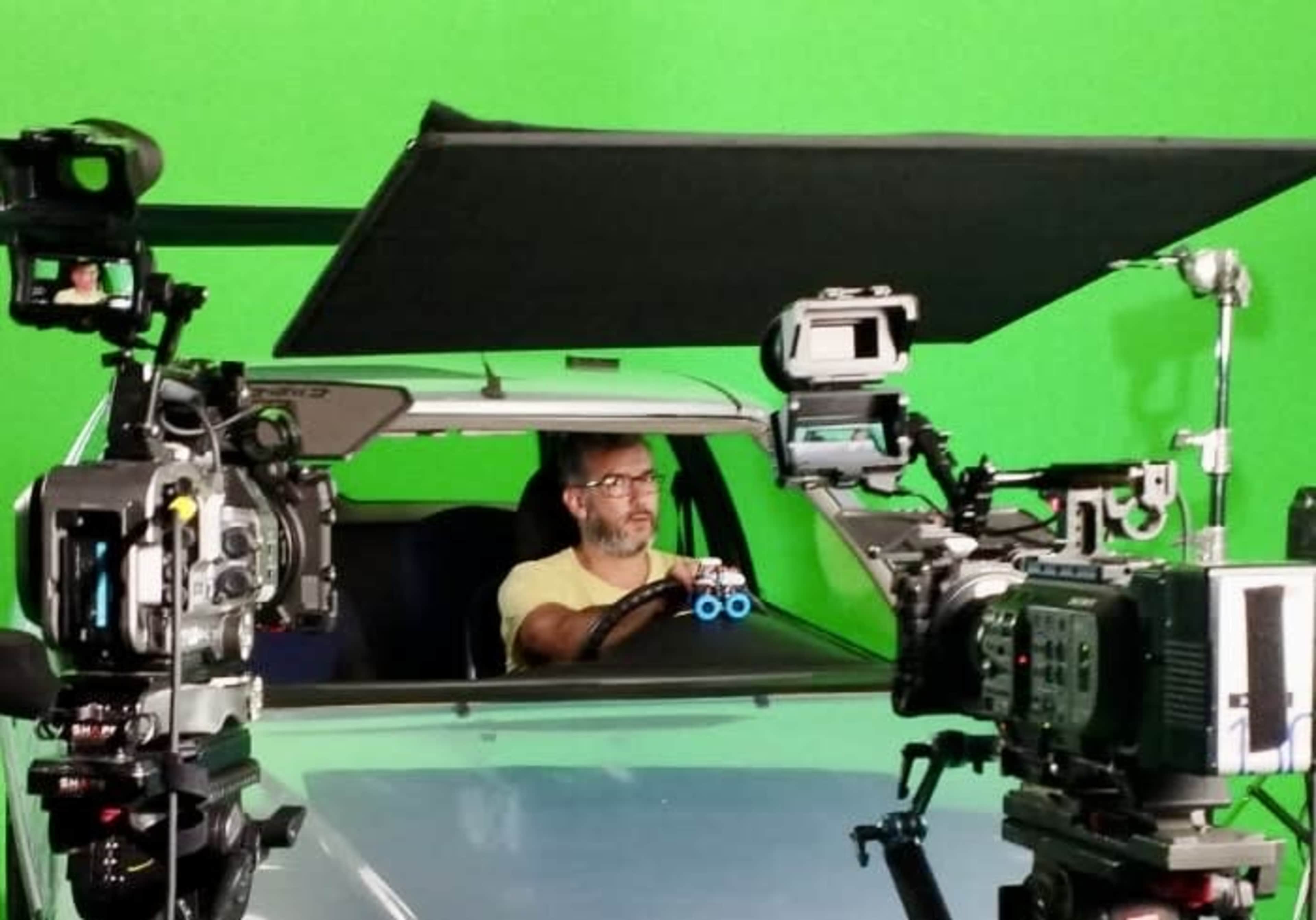 A man is seated in a vehicle with multiple cameras positioned around him against a green screen backdrop.