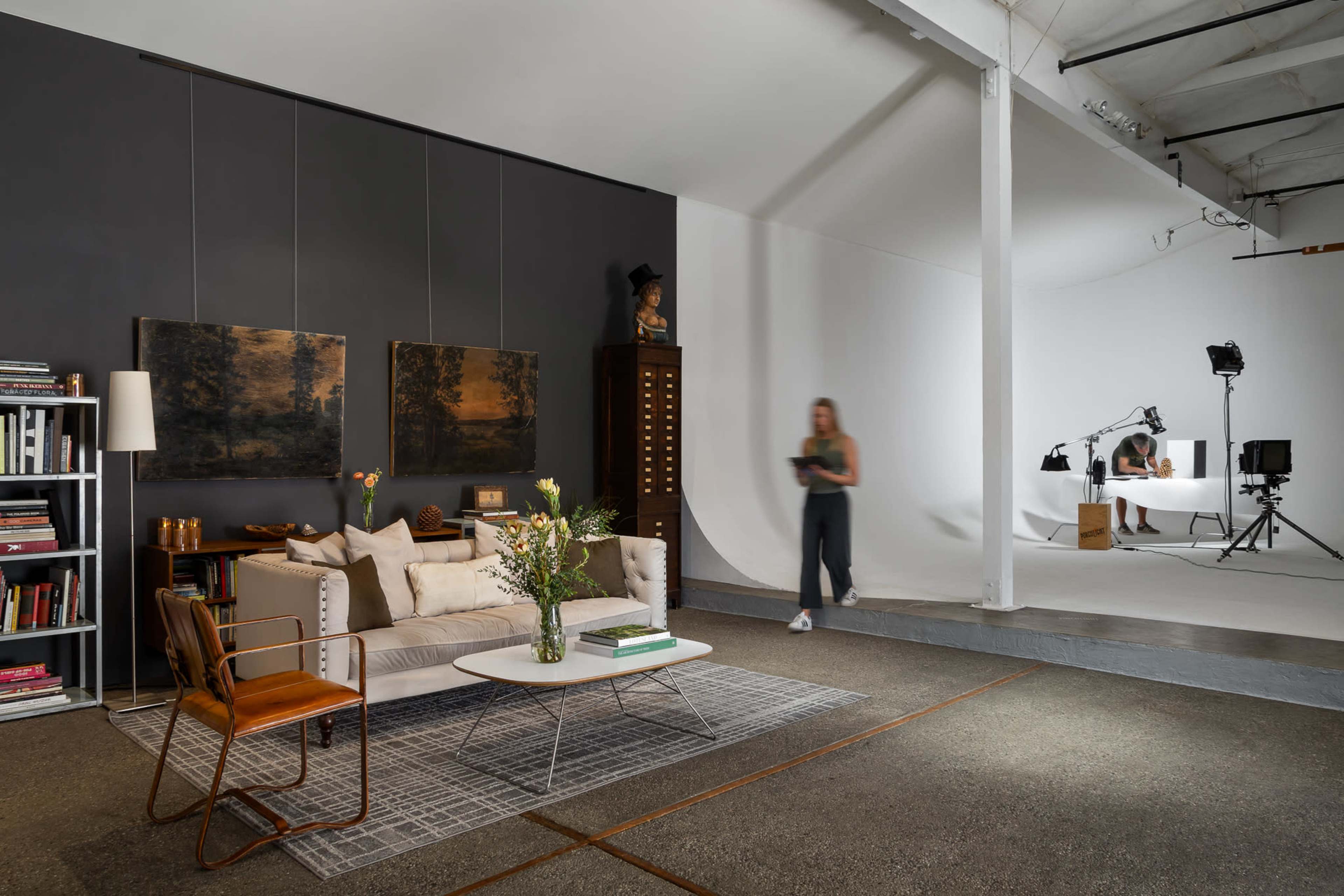A modern studio space features a stylish living area with a sofa, bookshelves, and decorative items, alongside a minimalist white backdrop and a workspace for photography.