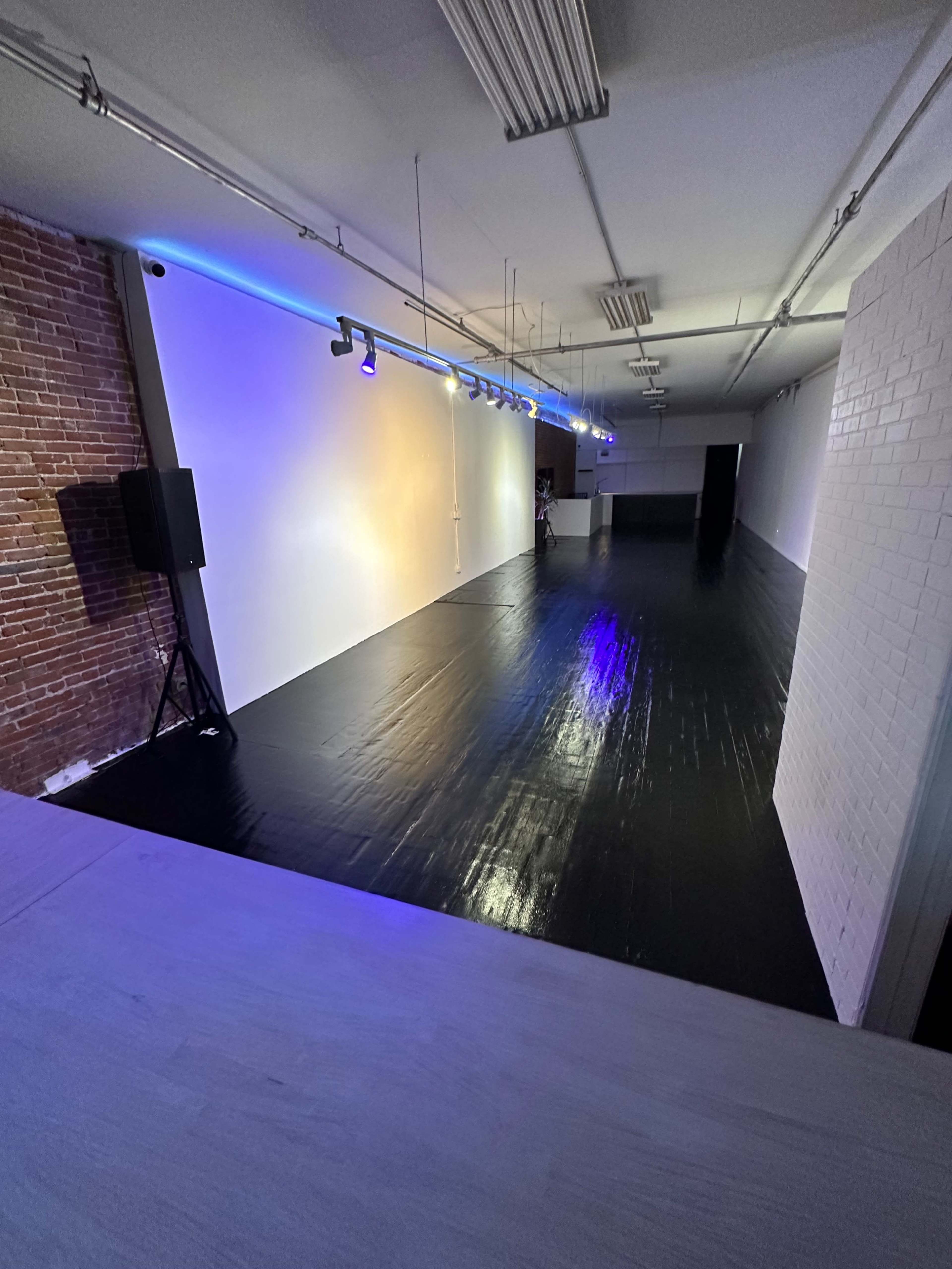 A spacious, empty room with a black floor, exposed brick wall, and lights illuminating one side.