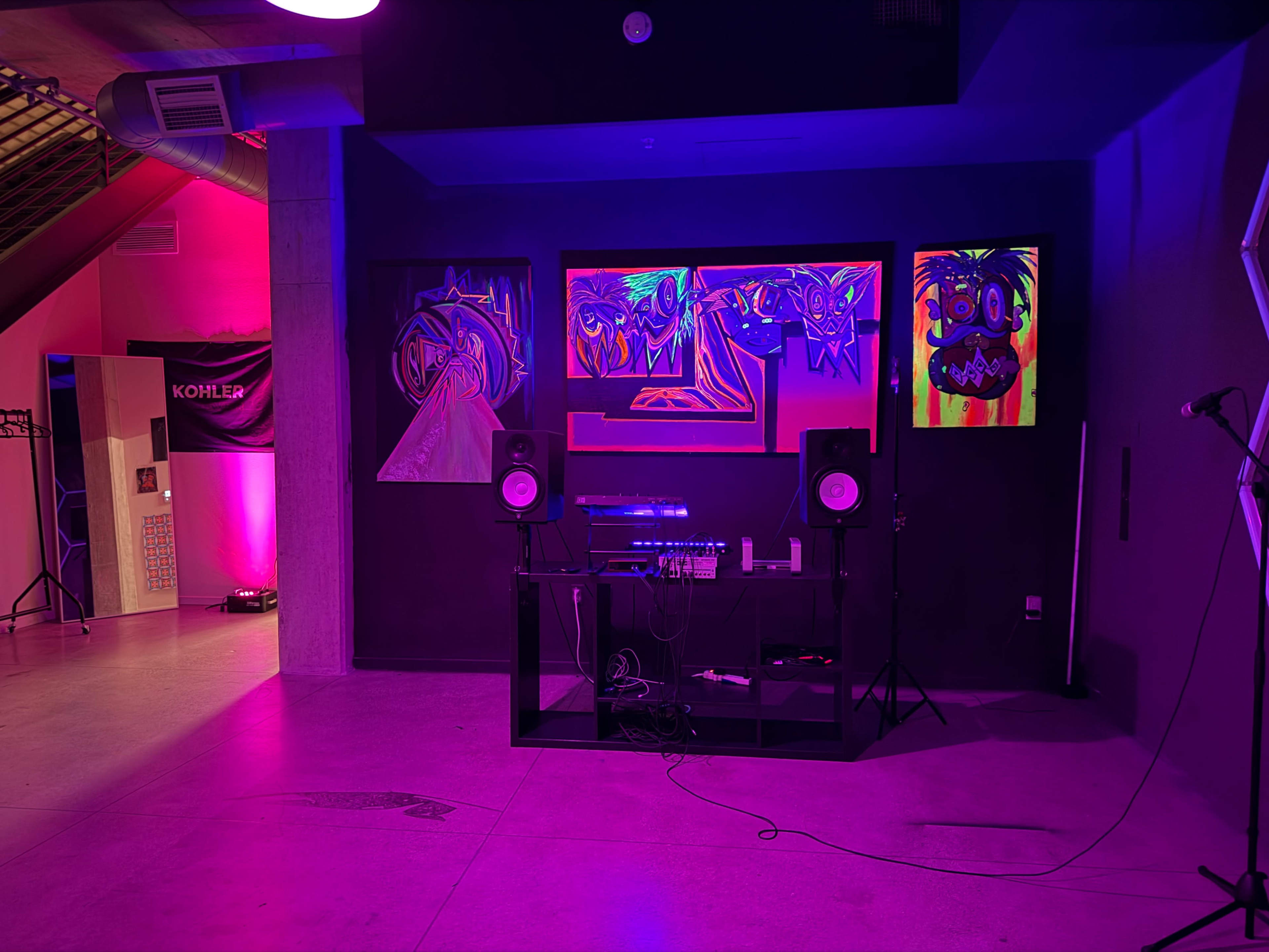 The image shows a dimly lit room featuring vibrant artwork on the walls, a sound system setup on a black table, and a microphone stand.