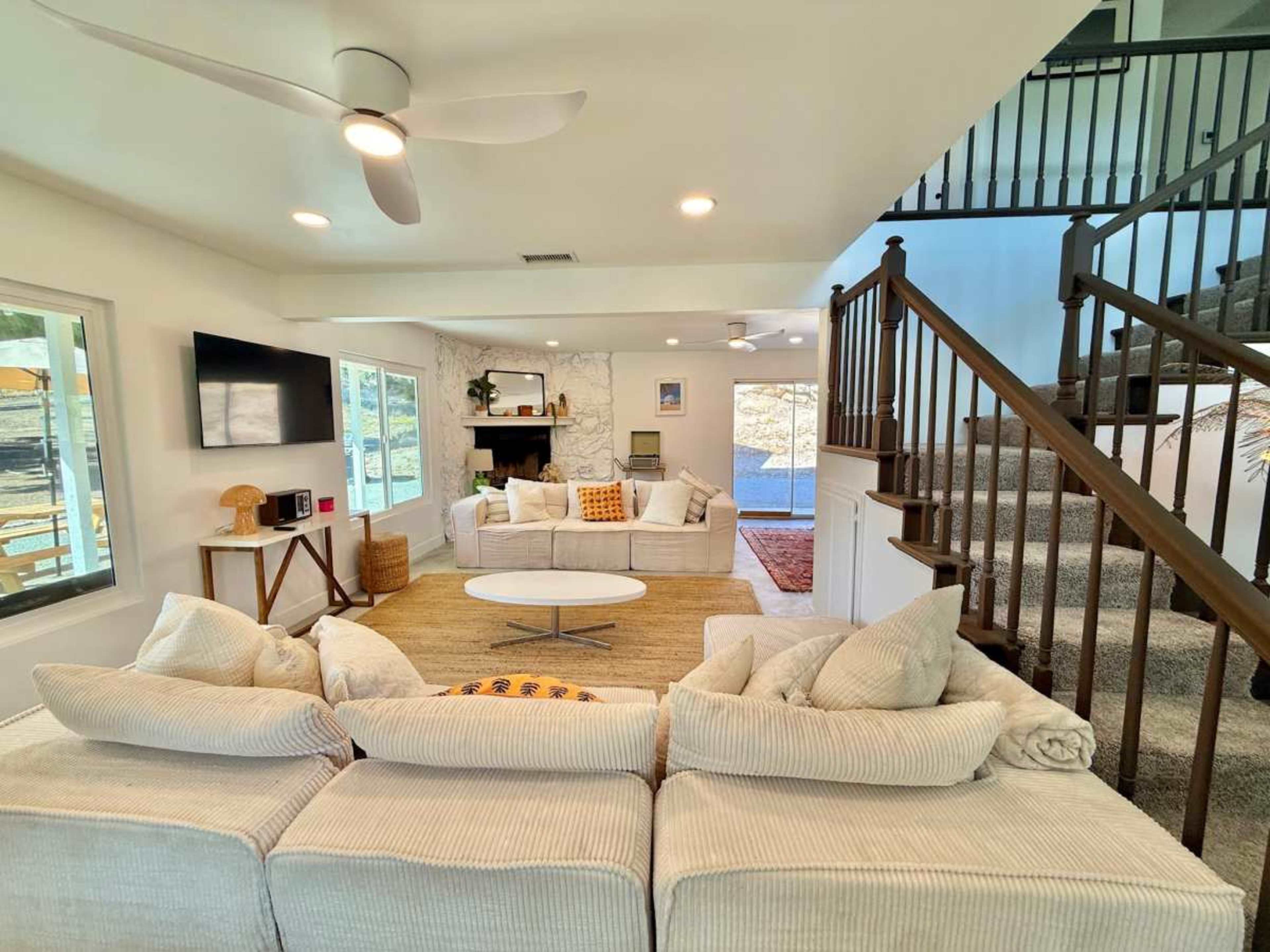 A bright living room features a large sectional sofa, a circular coffee table, and a staircase leading to the upper level, with a fireplace and large windows in the background.