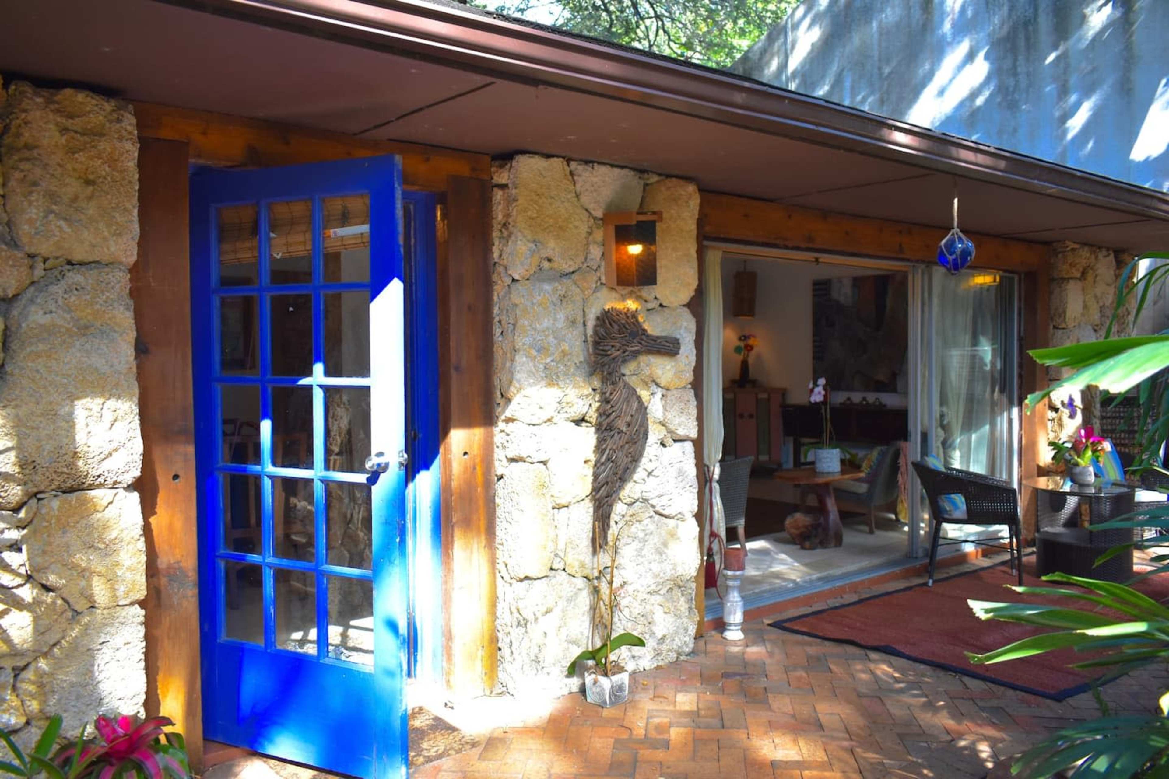 The image shows a stone-walled house with a blue door surrounded by greenery and a glimpse of indoor furnishings visible through an open entrance.