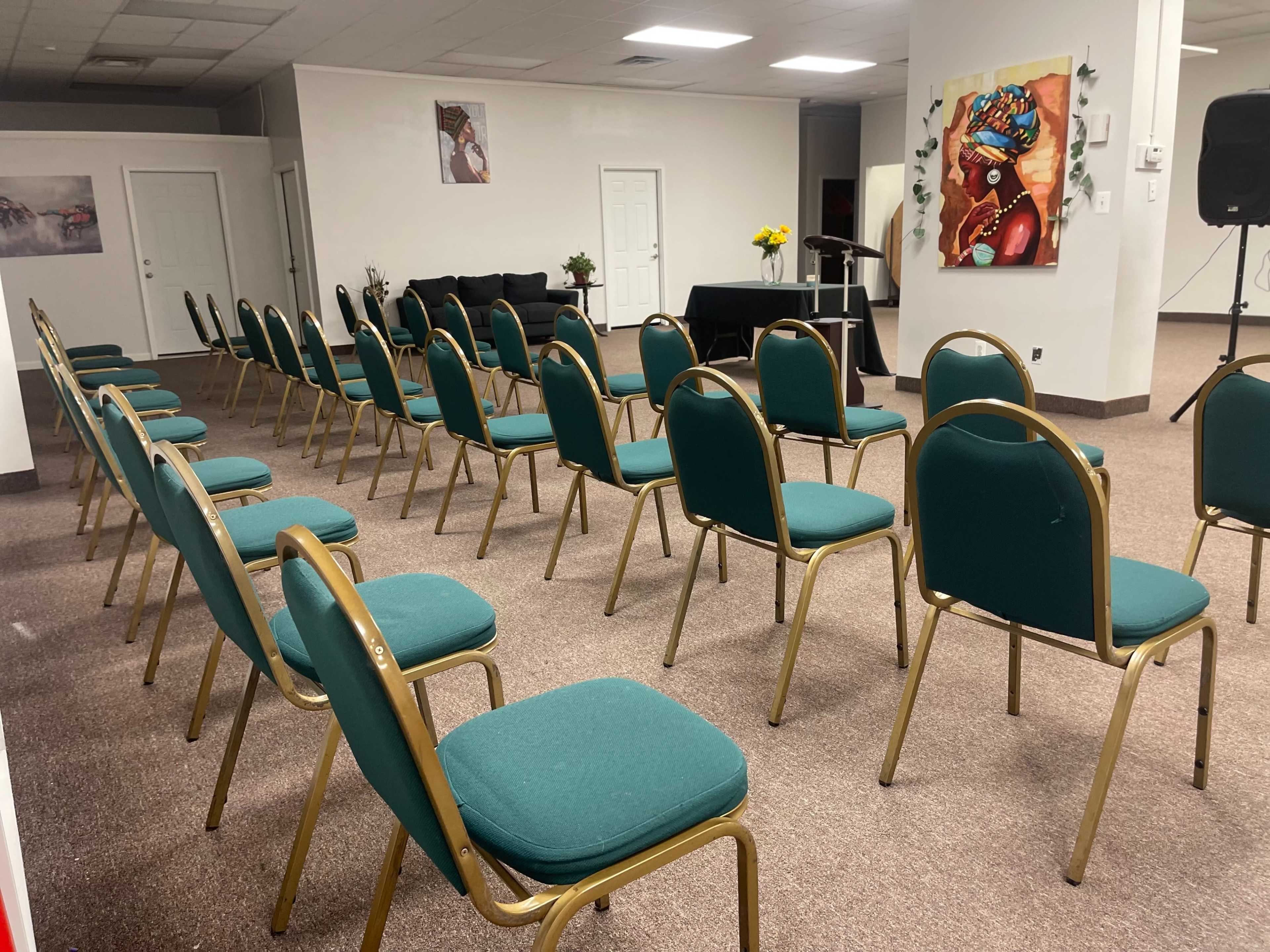 A room arranged with neatly aligned green chairs facing a small stage with a black table and flowers.