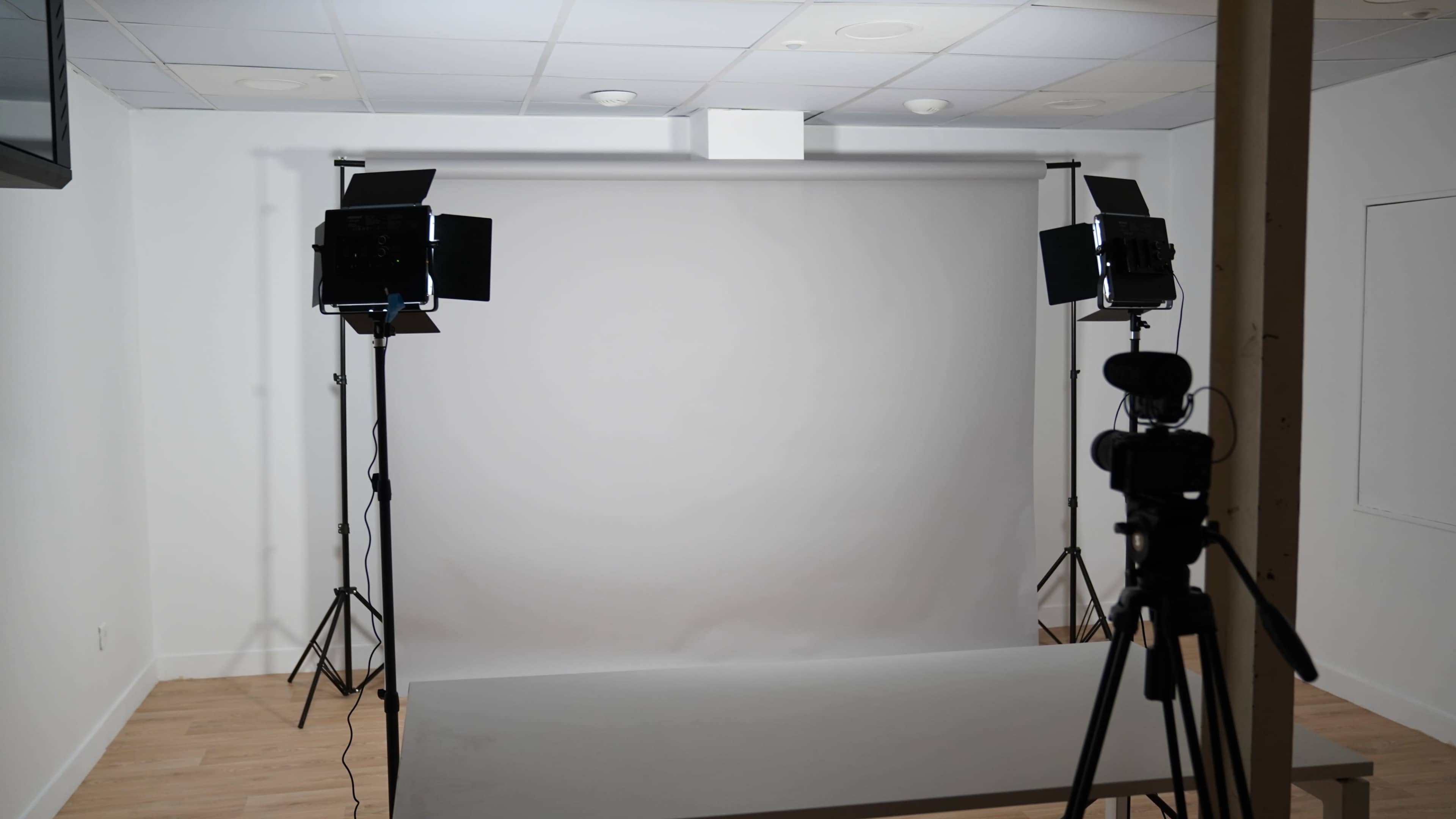 The image shows a minimalist photography studio featuring a grey backdrop, two softbox lights on stands, and a camera set on a tripod.