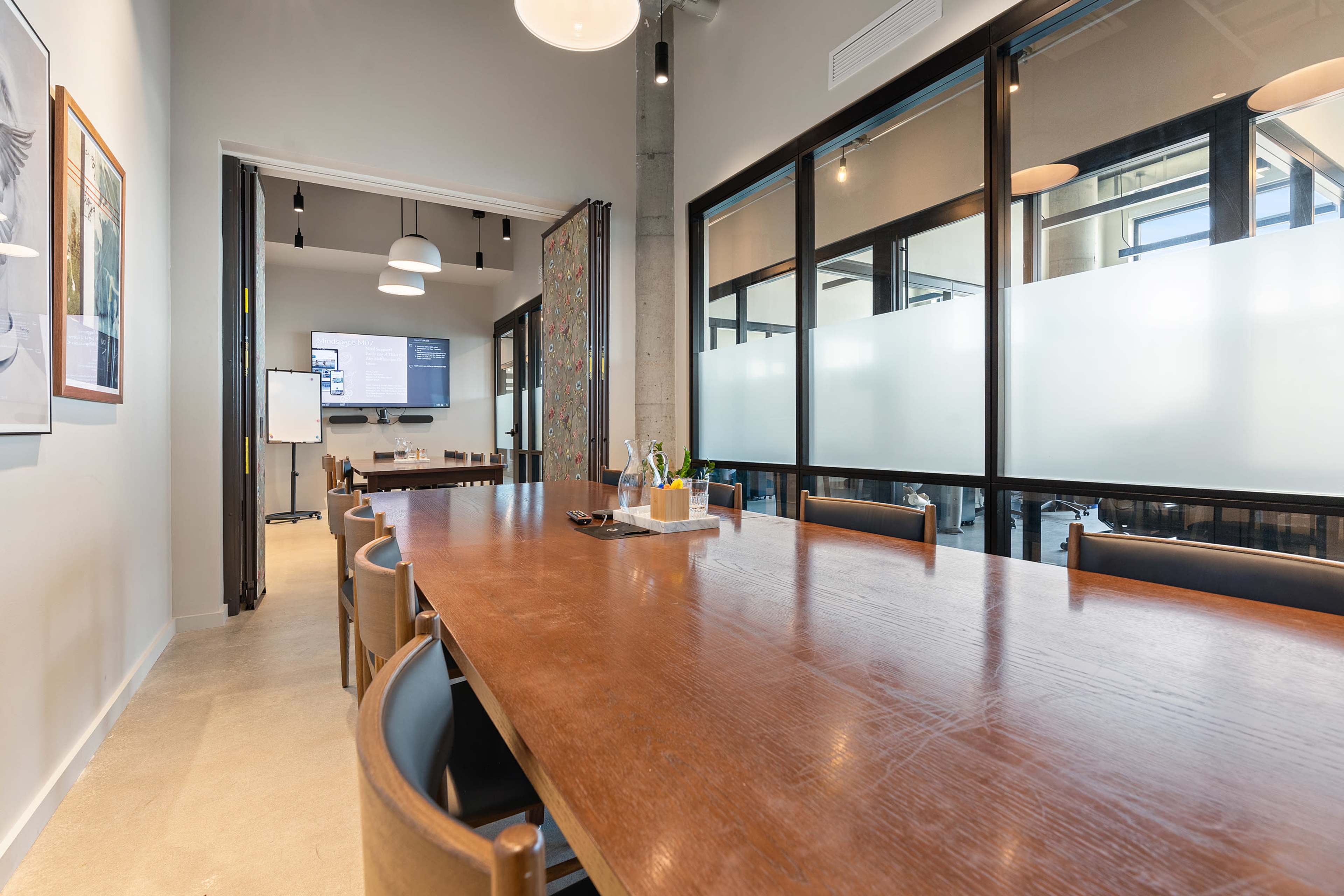A modern conference room with a long wooden table, sleek chairs, and a large screen displaying information on one wall.