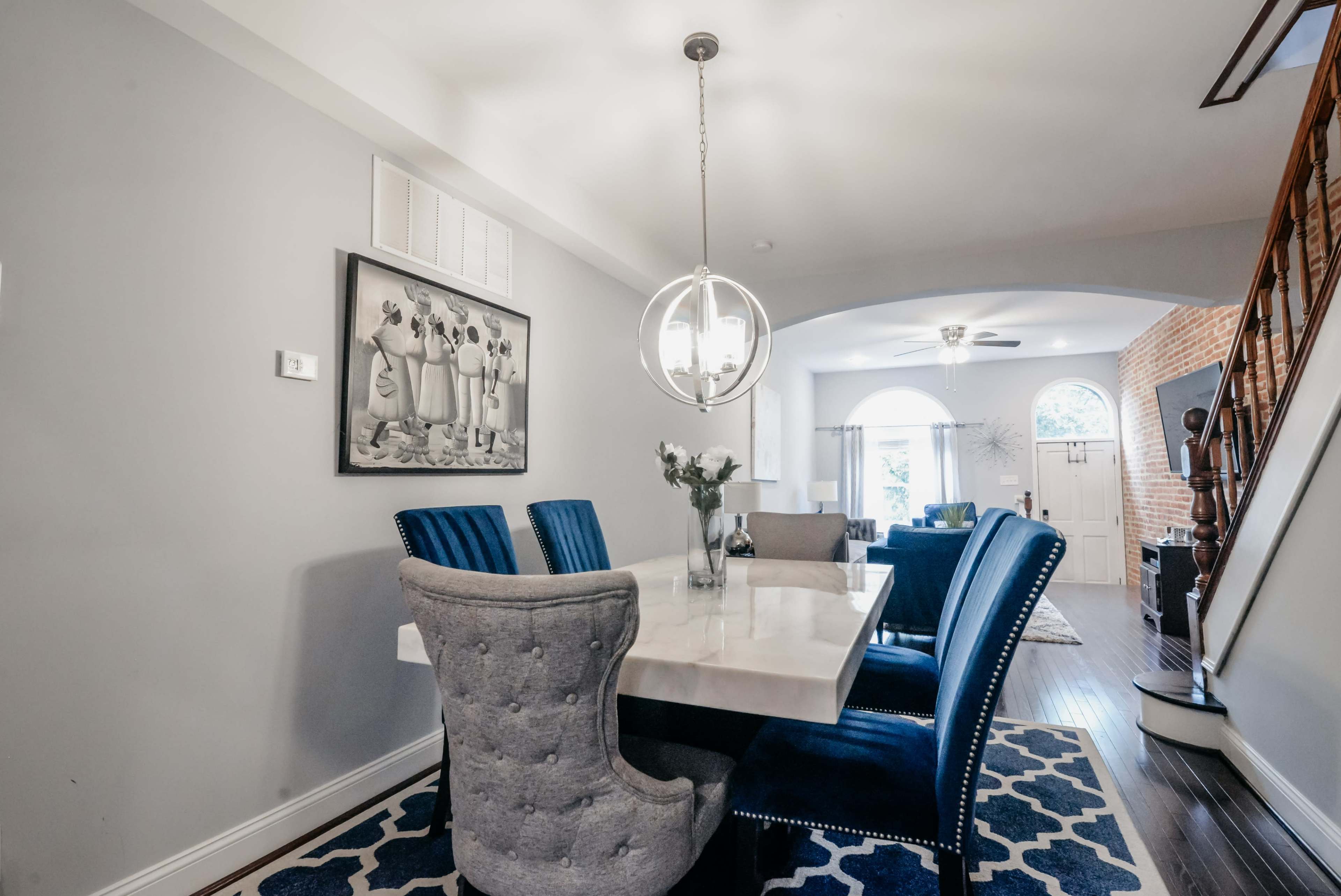 A dining room features a large marble table surrounded by blue and gray chairs, with a large light fixture overhead and a window in the background.