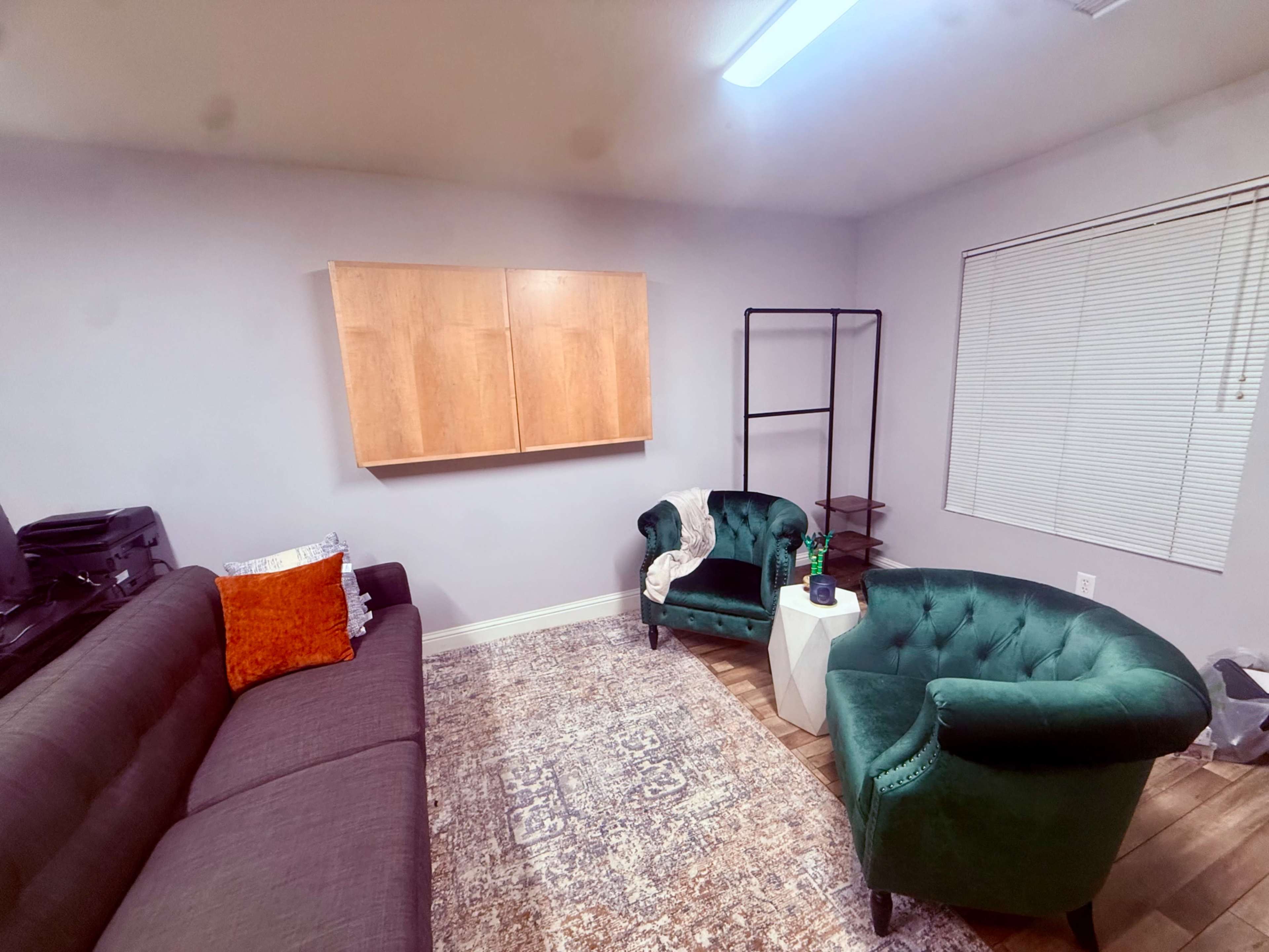 The image shows a cozy room with a dark gray sofa, two green chairs, a white side table, a wooden storage unit on the wall, and a window with blinds.