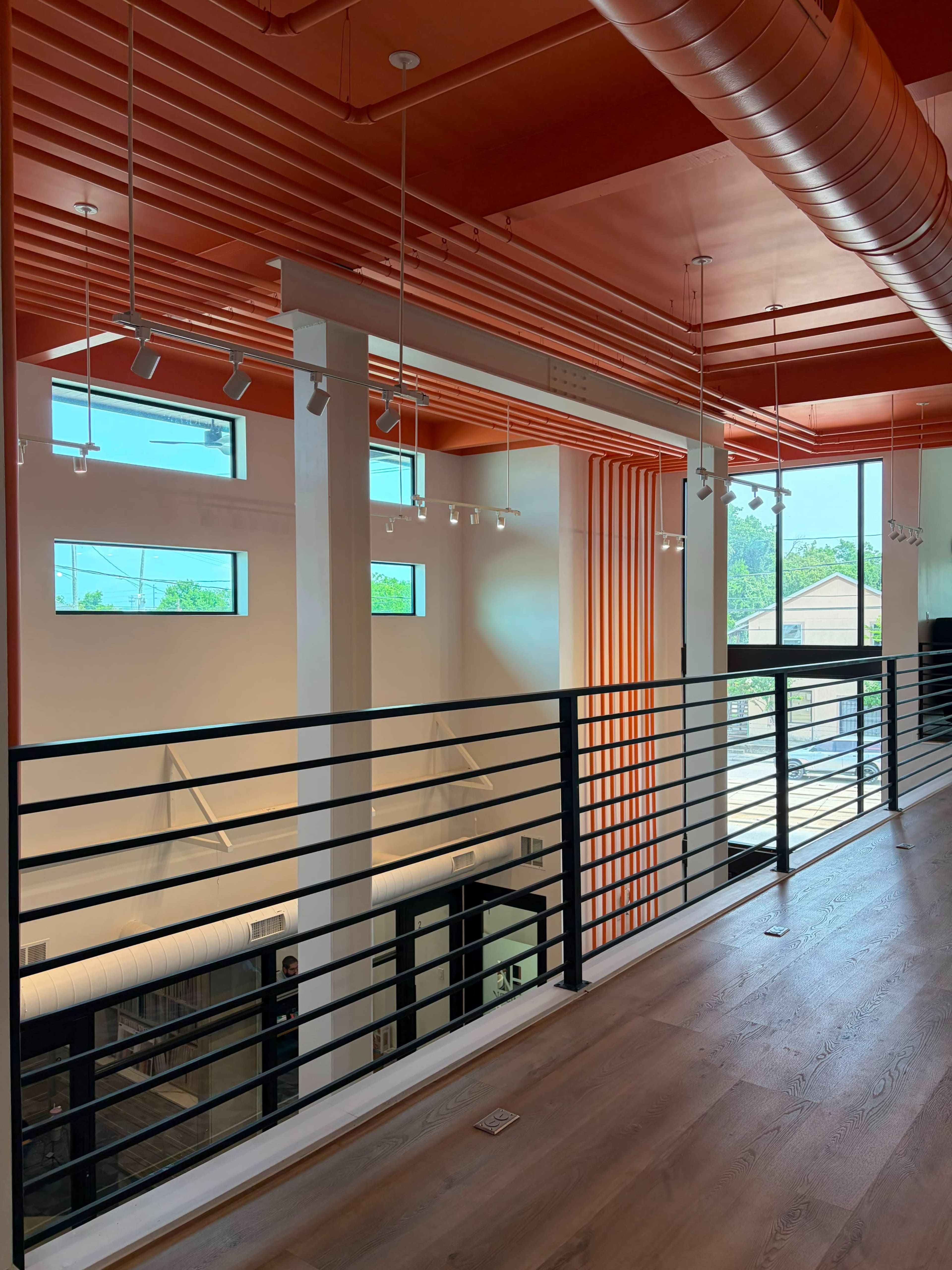The image shows a modern interior of a building, featuring a loft area with a black railing, orange ceiling, and large windows allowing natural light to enter the space.