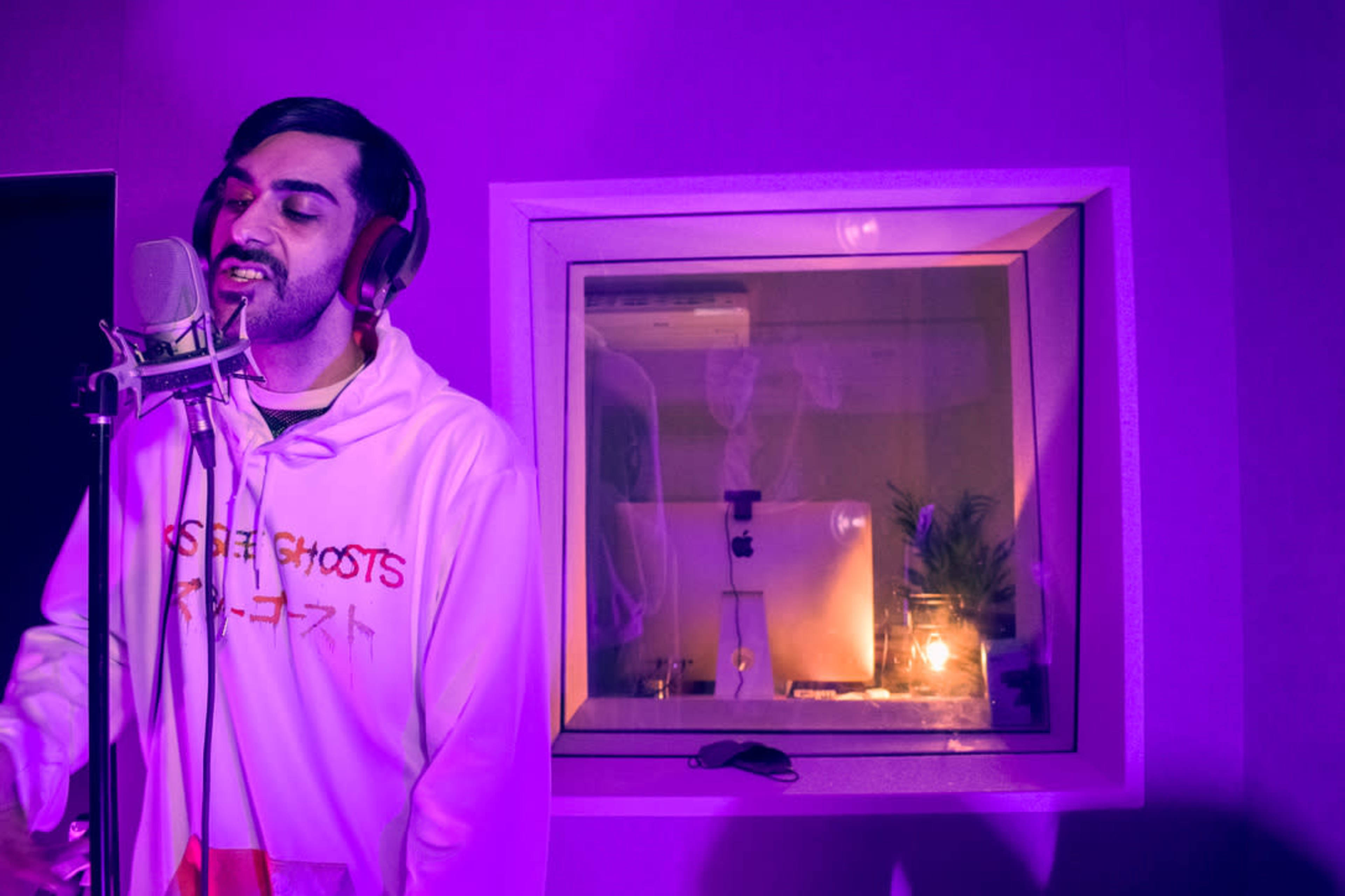 A man wearing a white hoodie sings into a microphone in a recording studio illuminated by purple lighting, with a reflection visible in a glass window.