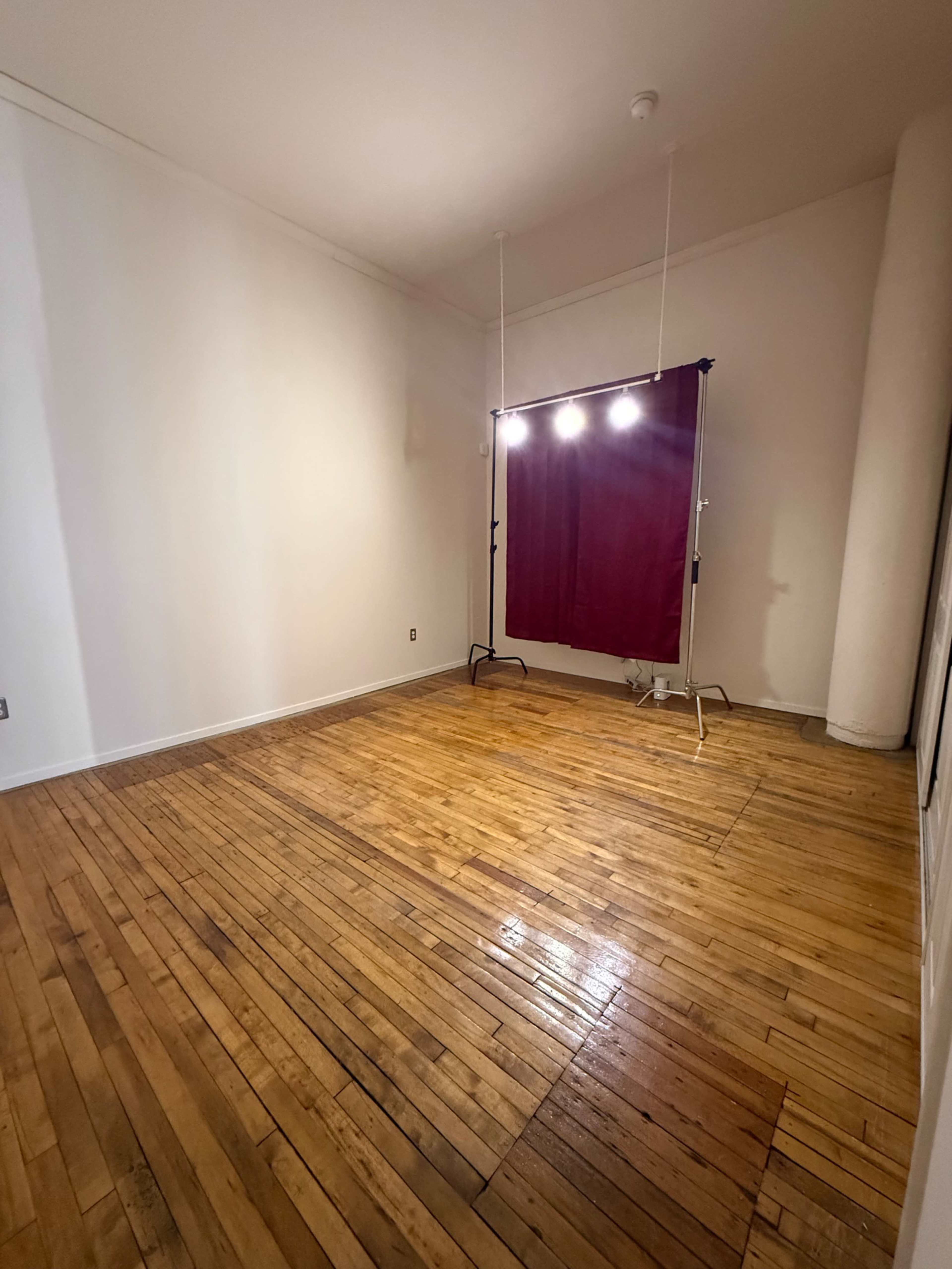 The image shows a spacious, empty room with polished wooden floors and a curtain backdrop illuminated by overhead lights.