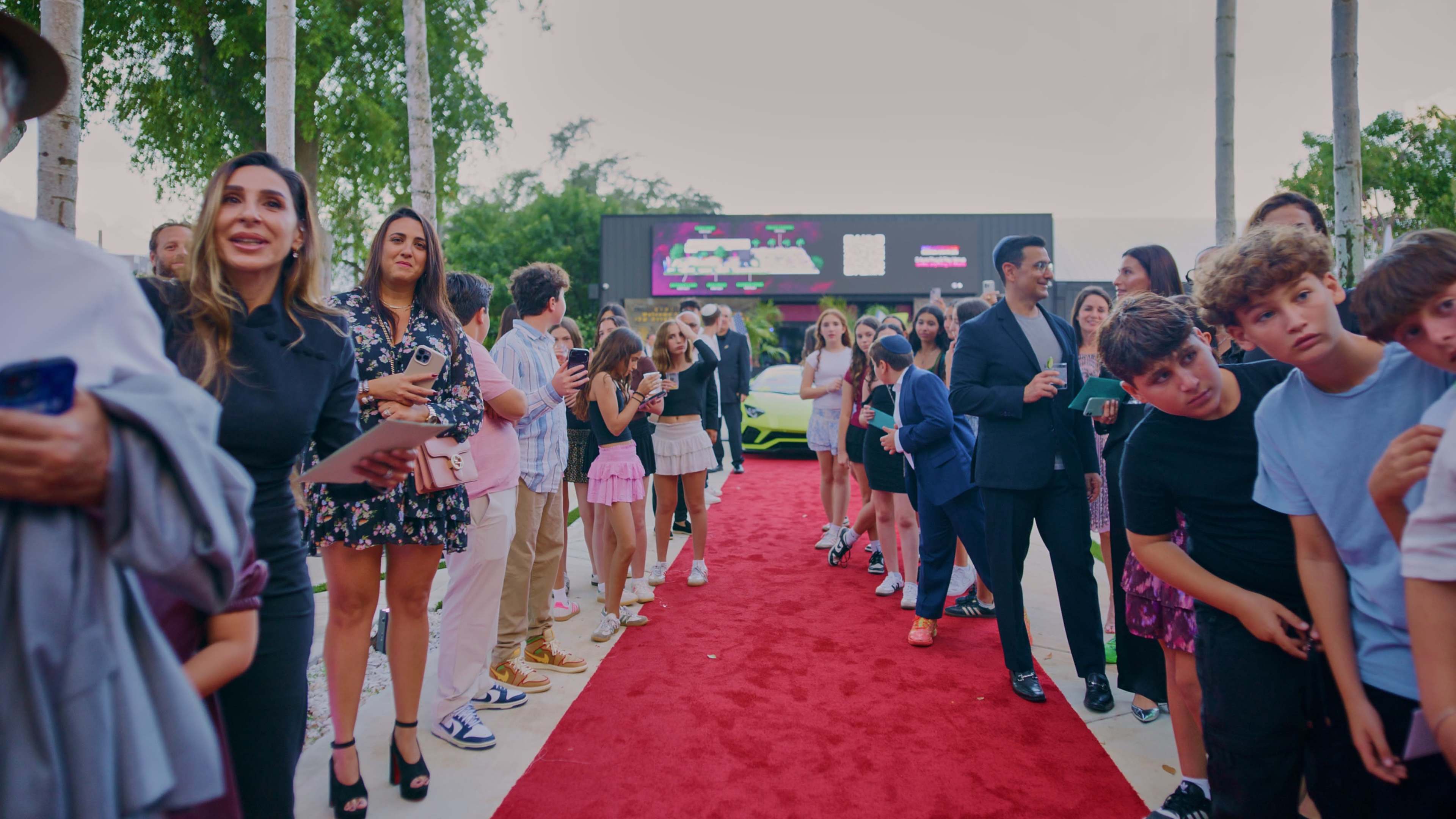 A crowd gathers along a red carpet outside a venue, with attendees dressed in various outfits and a luxury car visible in the background.