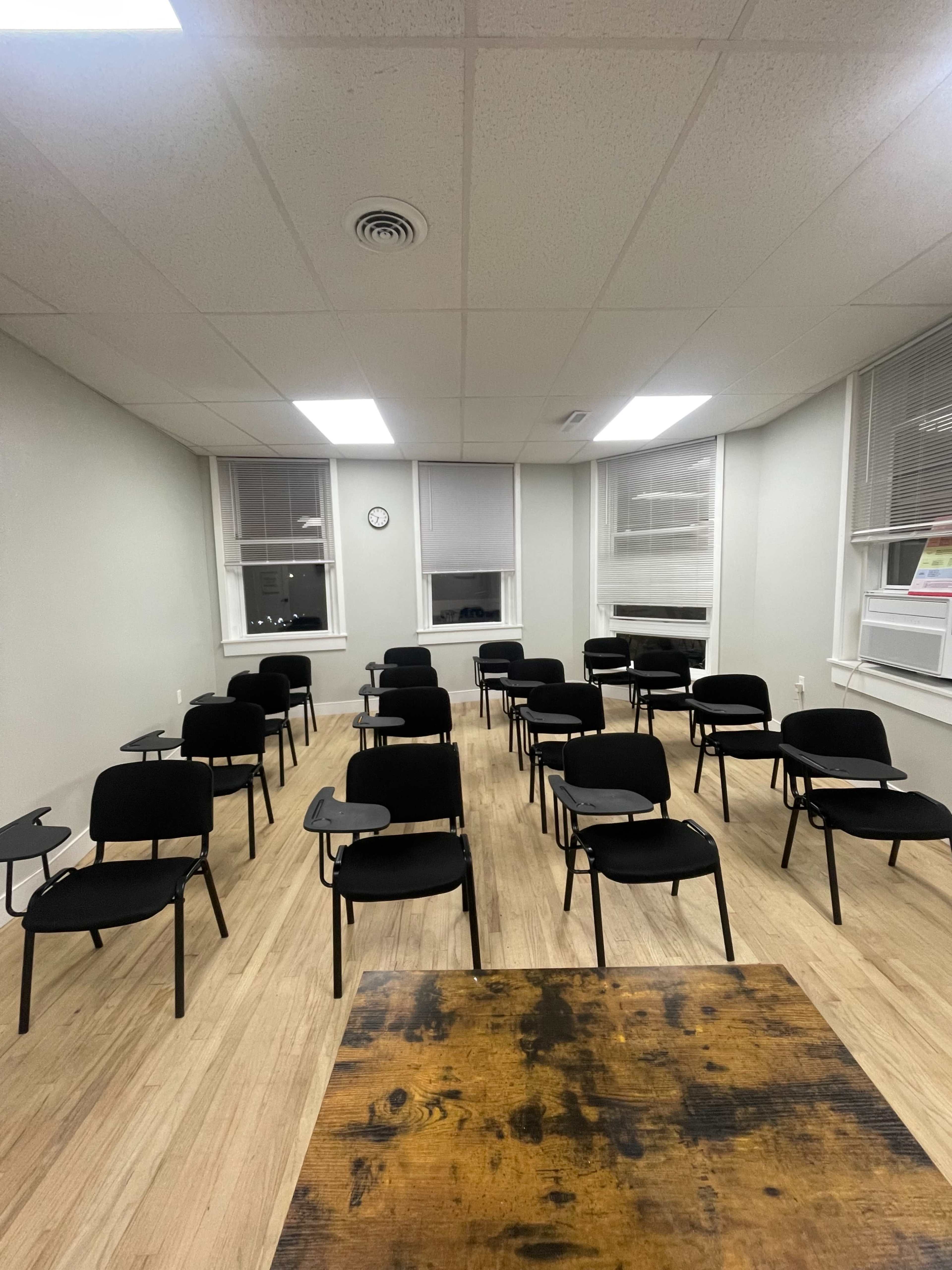 The image shows a classroom with several rows of black chairs equipped with small writing surfaces arranged around a wooden table.