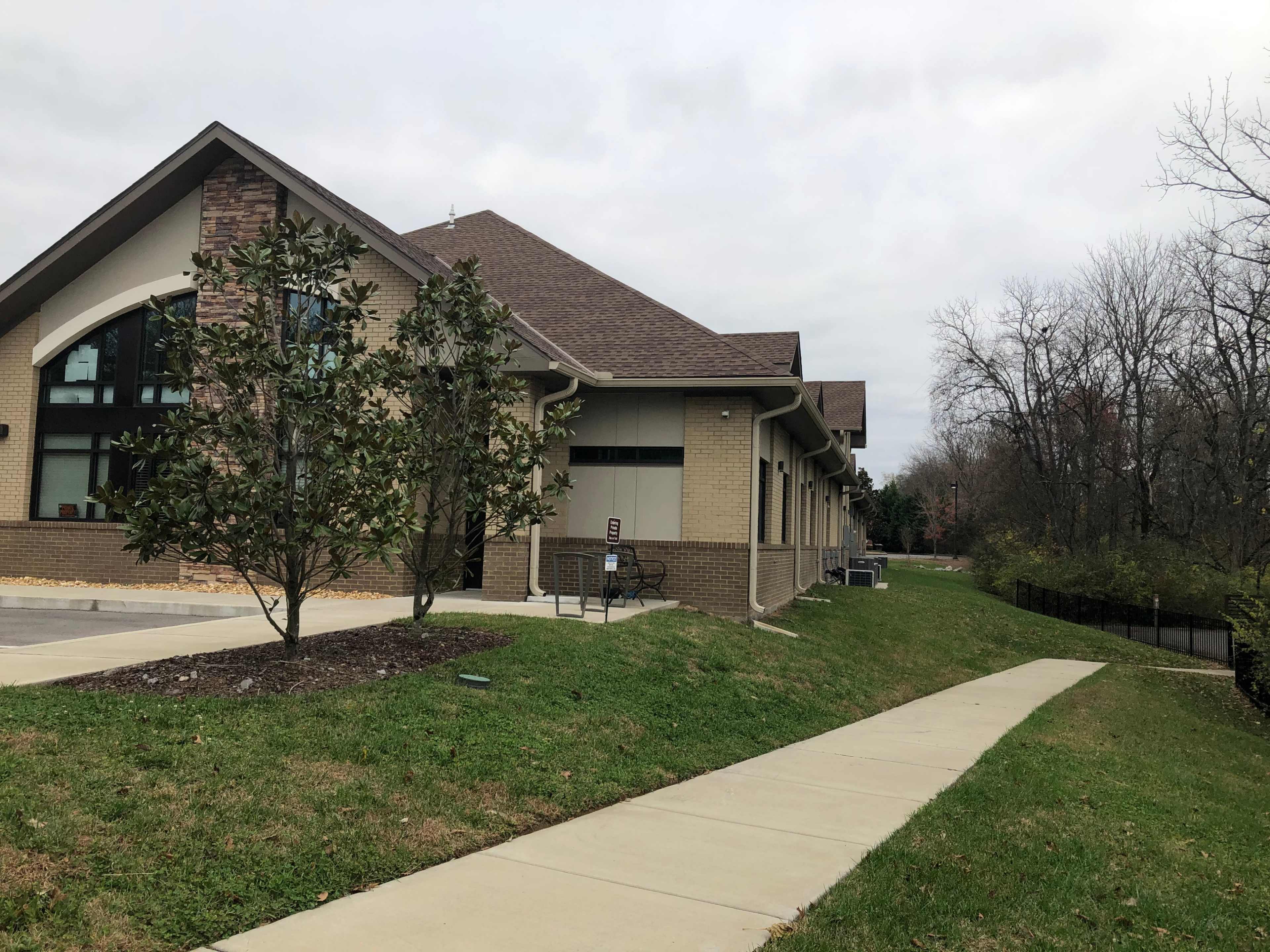 A modern beige building with a sloped roof is situated next to a paved pathway, surrounded by grass and a few trees.