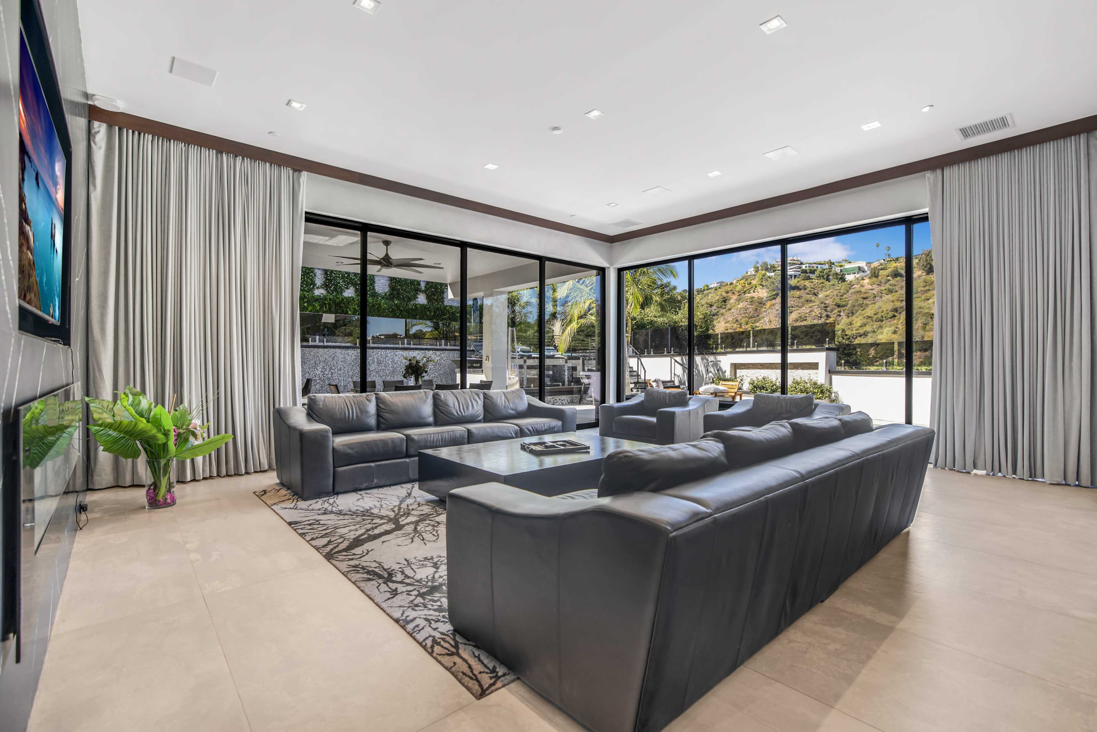 A modern living room features large windows overlooking a scenic outdoor area, with dark leather sofas arranged around a coffee table on a patterned rug.