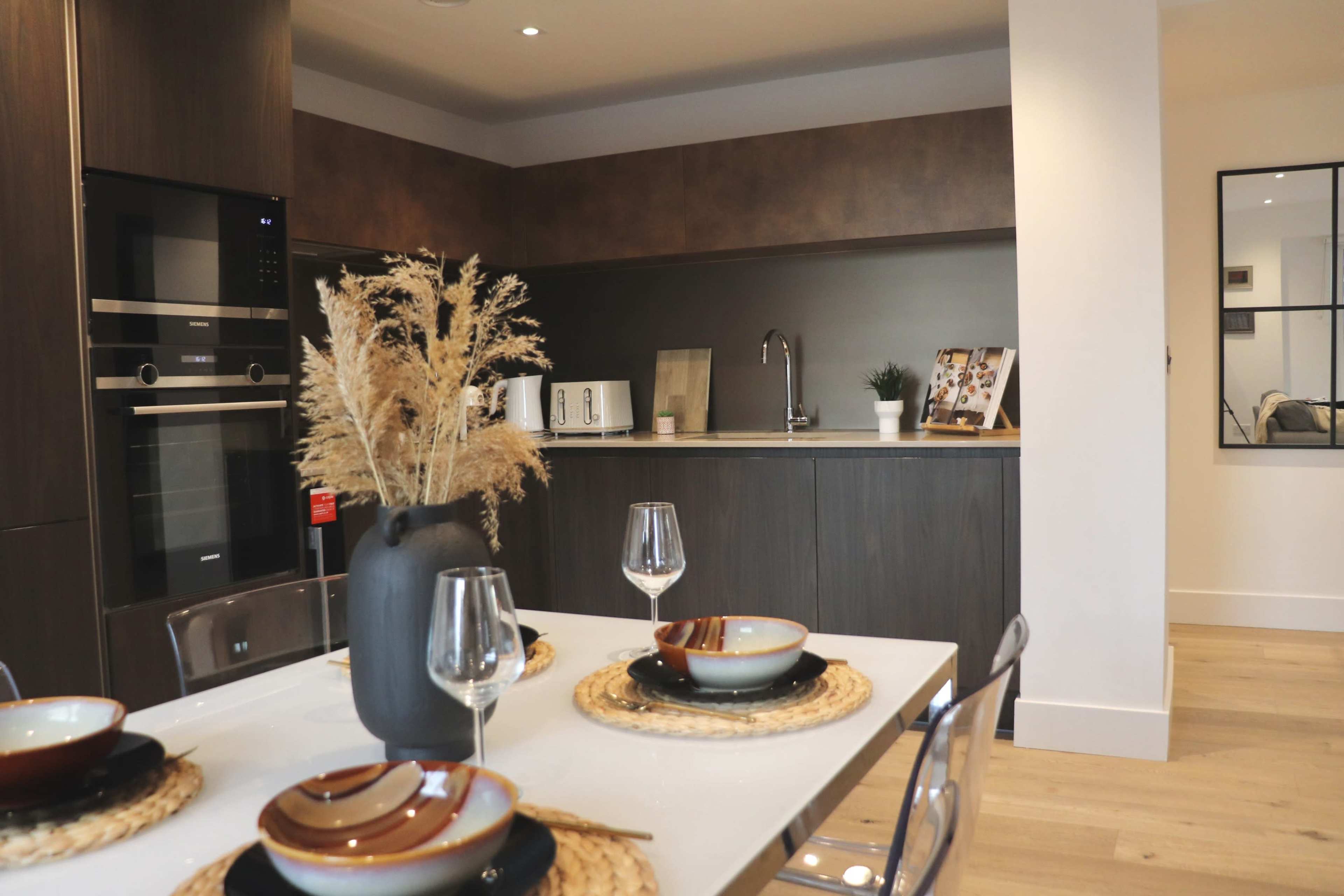 A modern kitchen and dining area features a table set with plates and glasses, alongside a minimalist kitchen equipped with appliances and decorative elements.