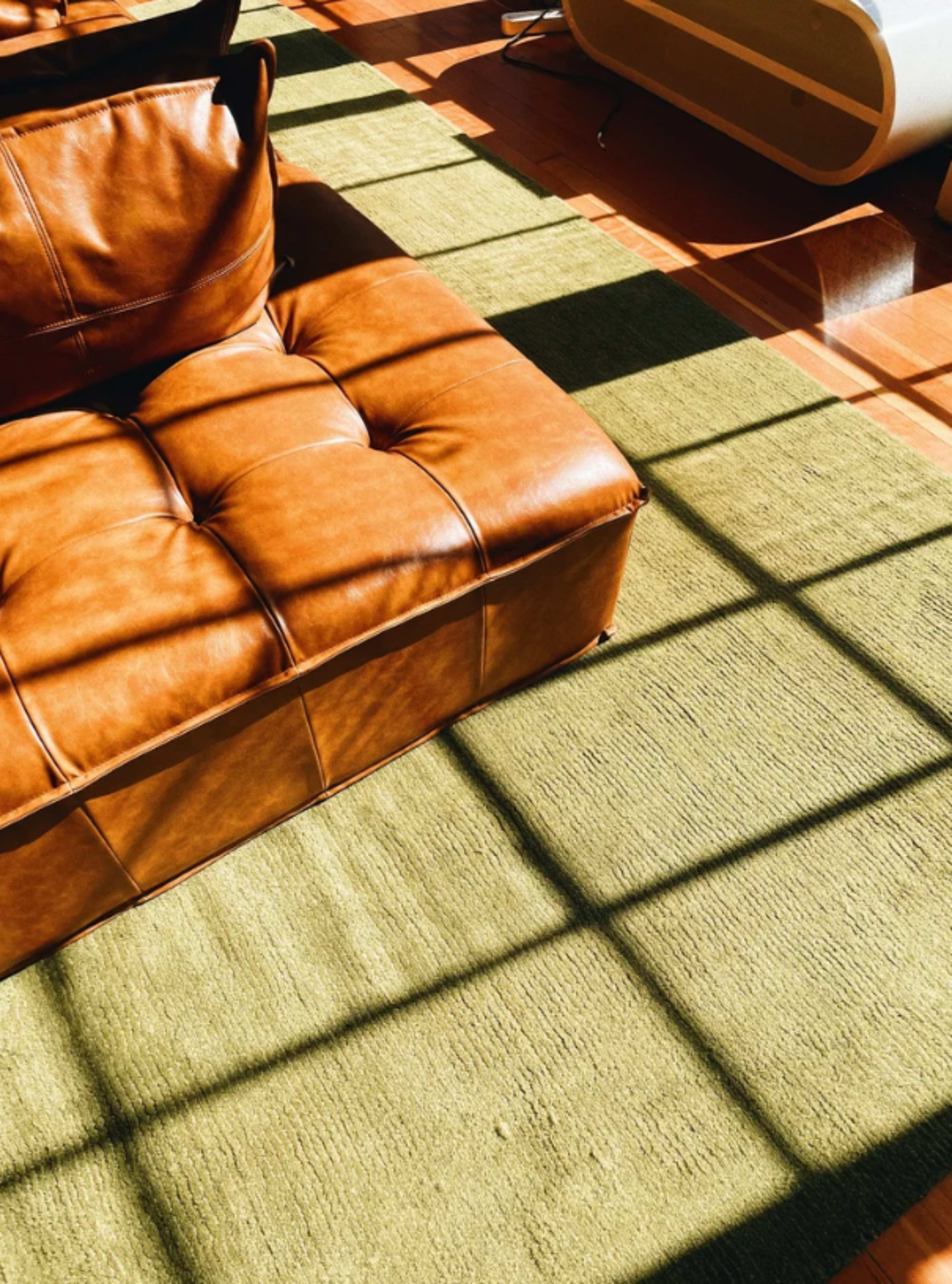 A brown leather tufted ottoman sits on a green rug, illuminated by sunlight creating patterned shadows on the floor.