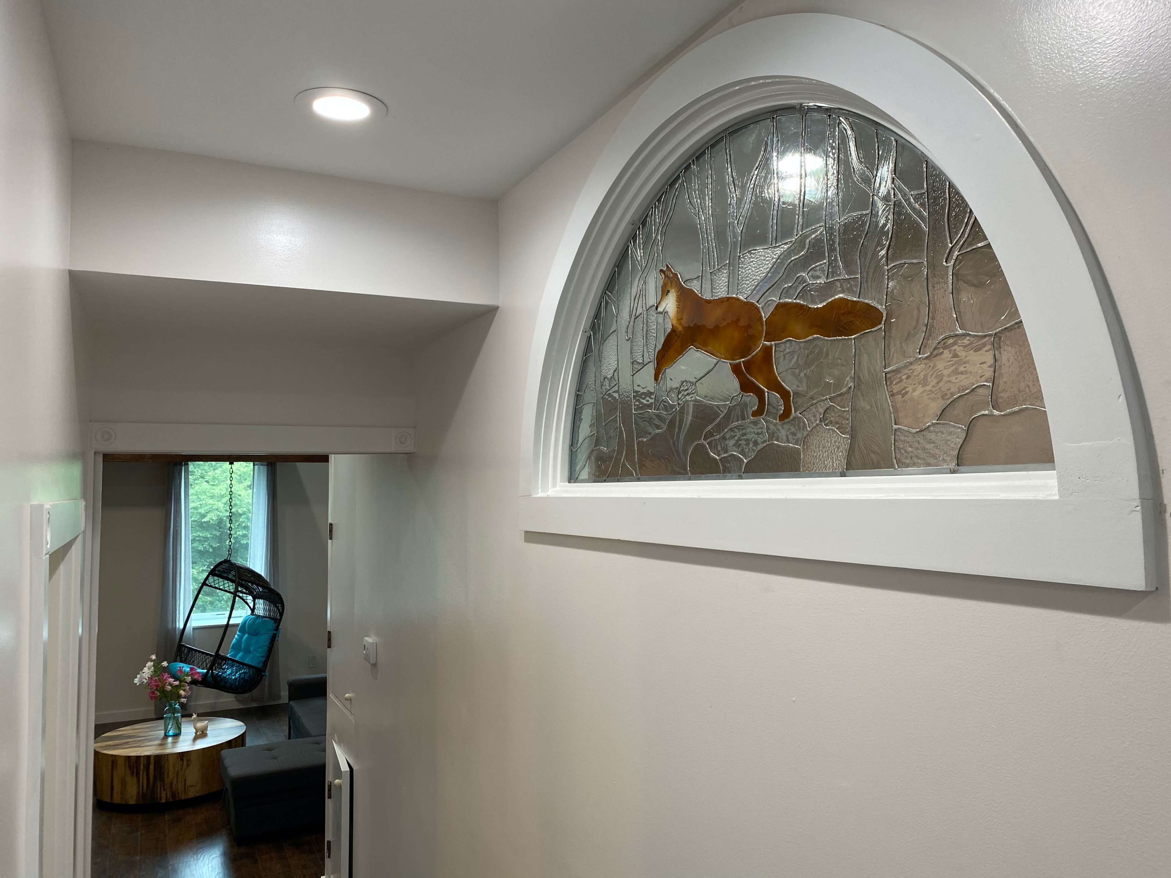 A hallway features an arched stained glass window depicting a fox, with a cozy seating area visible in the background.