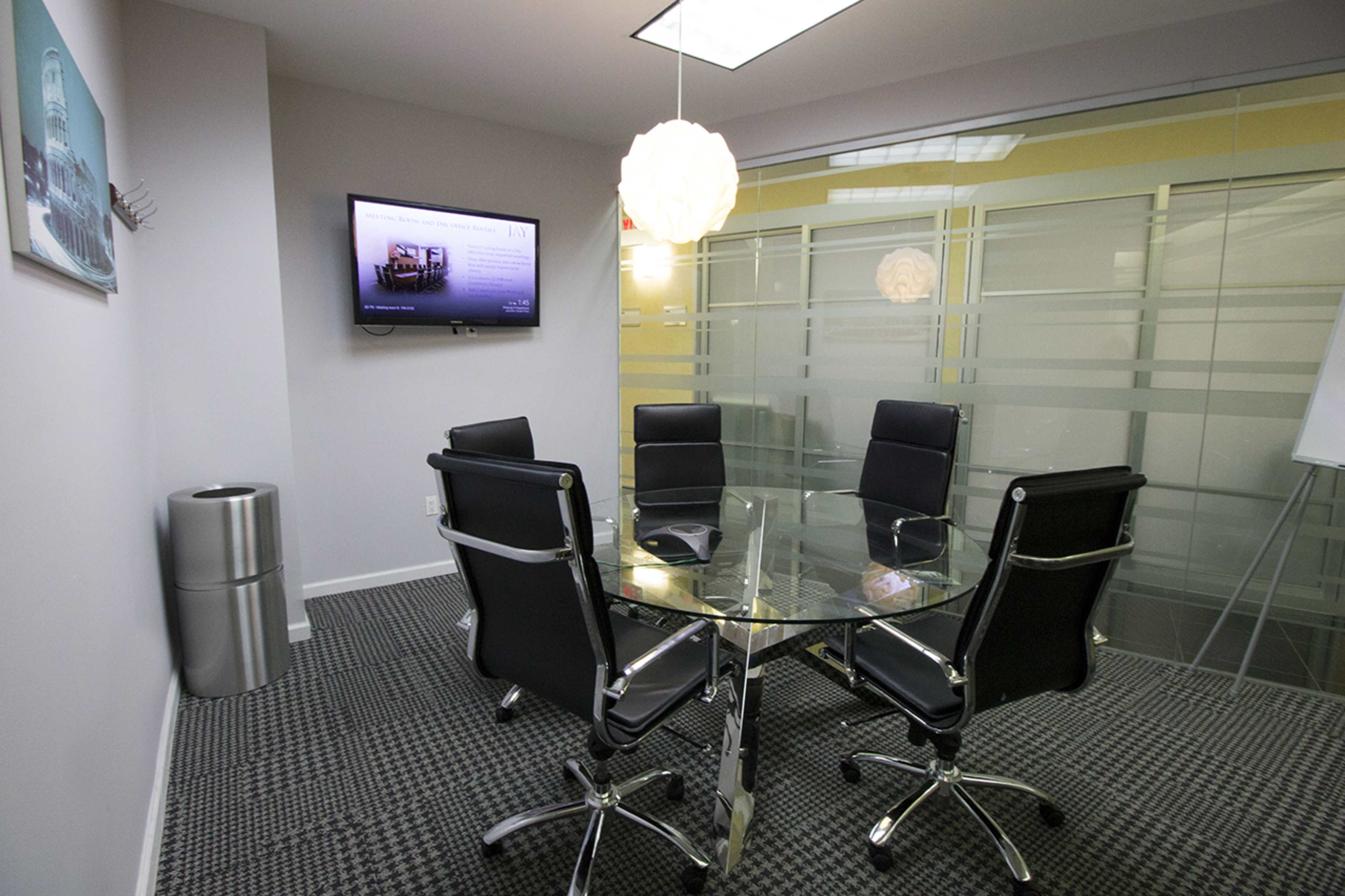 A modern conference room features a glass table surrounded by black chairs, with a television mounted on the wall and a light fixture above.