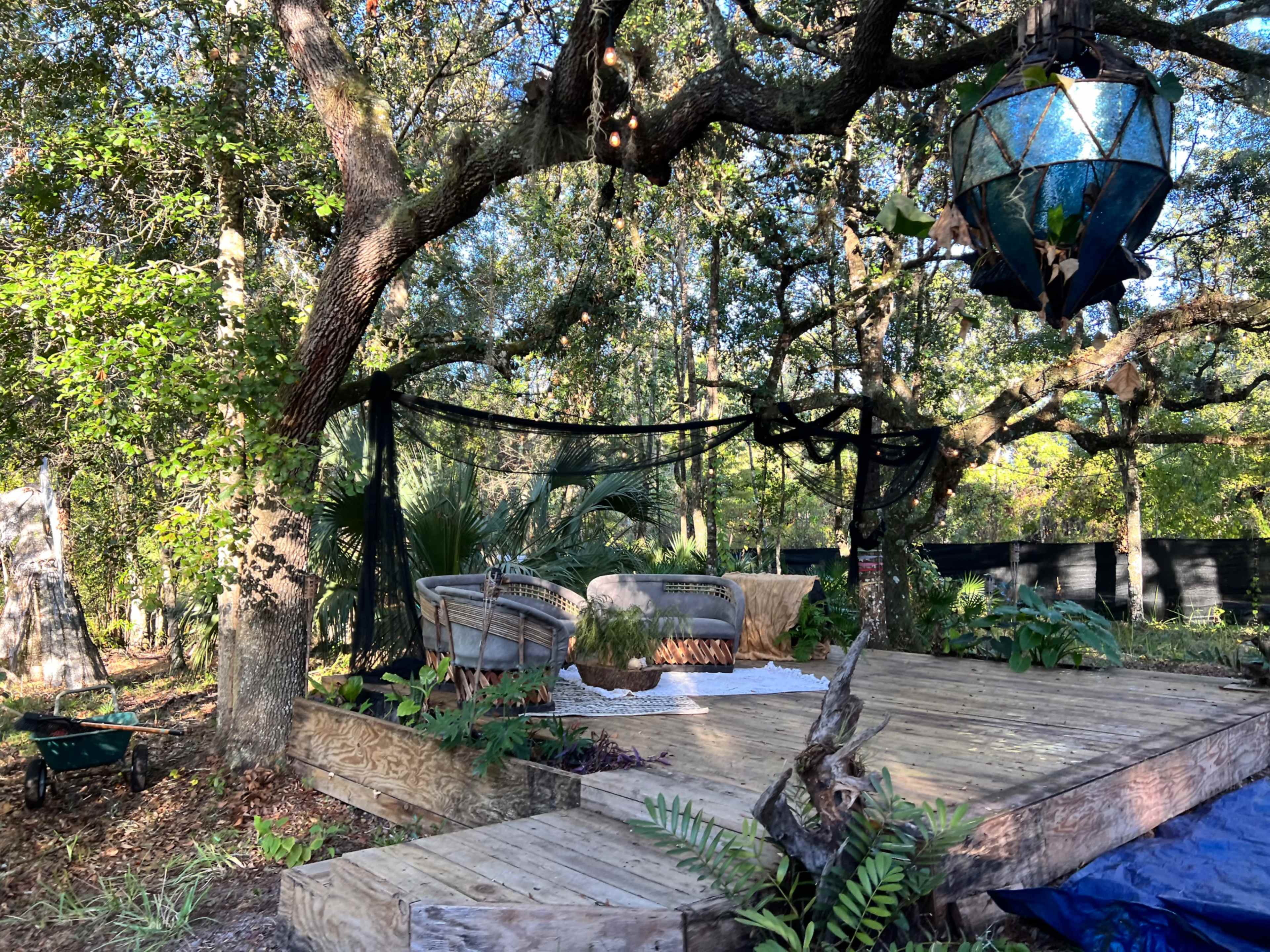 A wooden deck under an oak tree features two wicker chairs, a hanging lantern, and various plants around it.