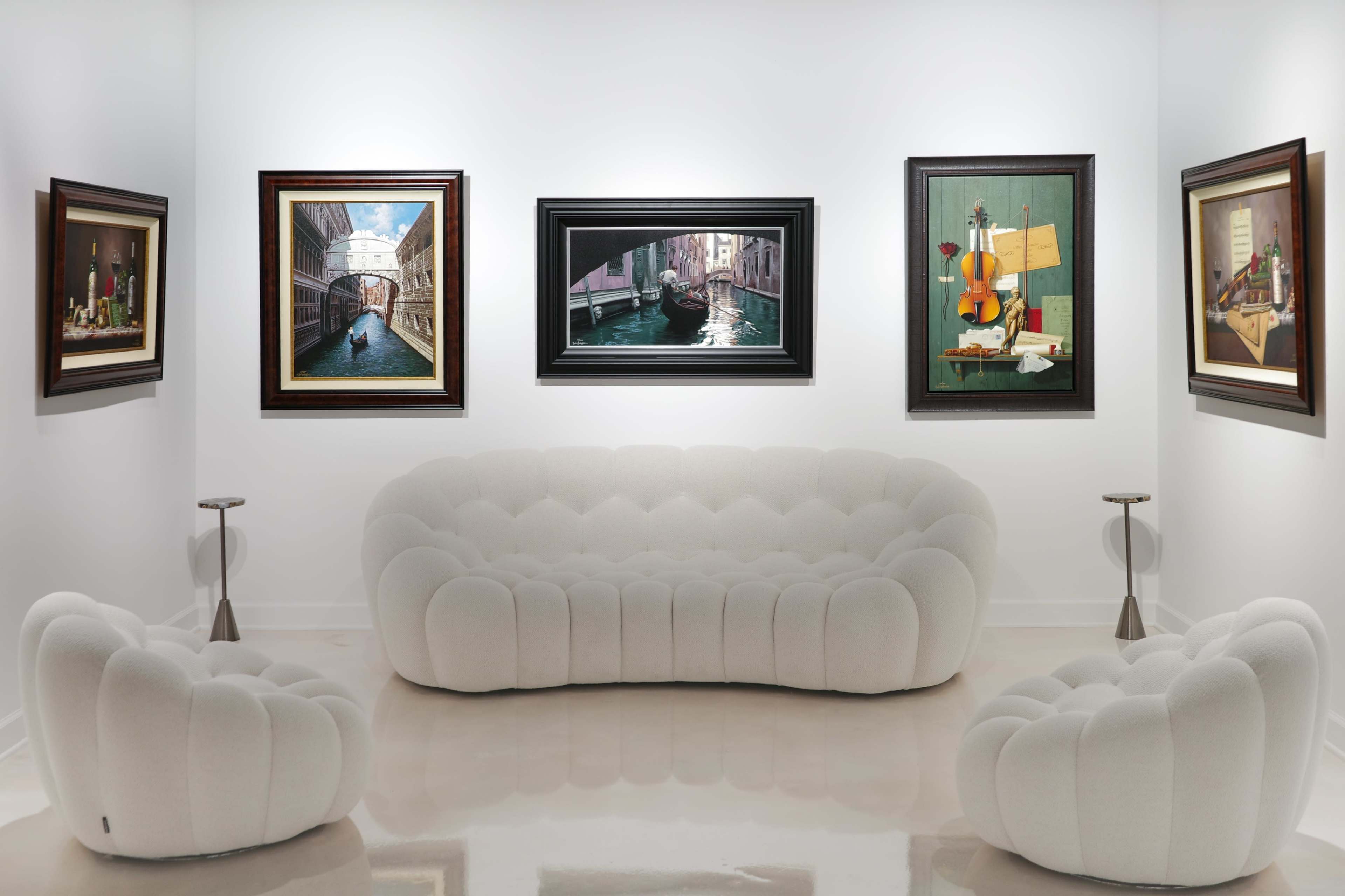 A modern art gallery features a white, rounded sofa and two matching chairs, surrounded by framed paintings on the walls.