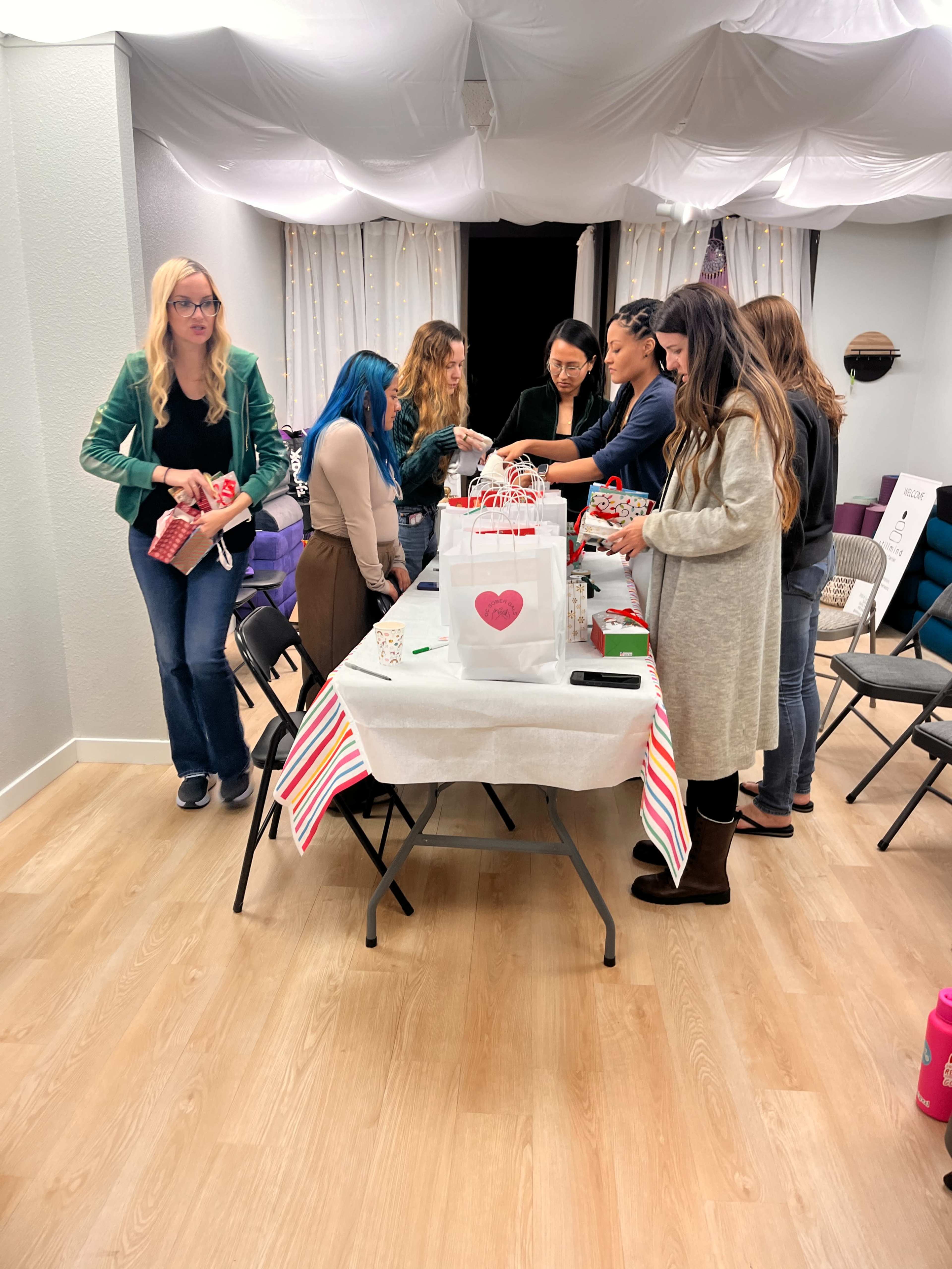 A group of women is gathered around a table, wrapping gifts while seated and standing in a well-lit room.